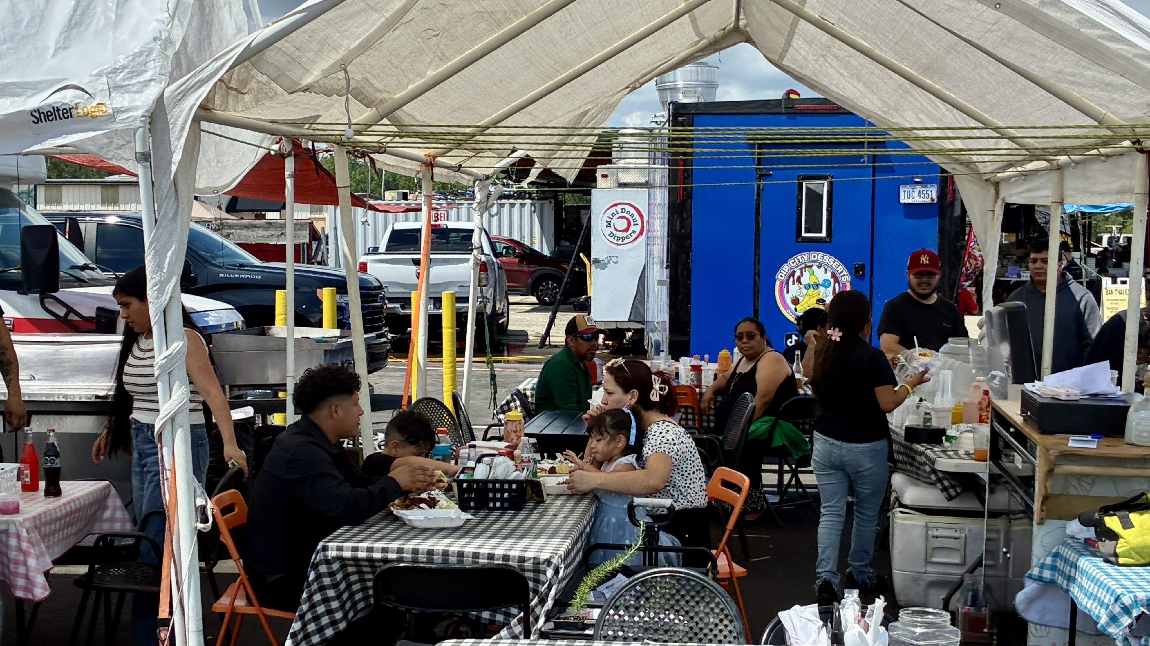 Busy outdoor dining under a white canopy with checkered tables; people eat and chat while condiment bottles sit on tables, and a blue food trailer with signs sits in the background.