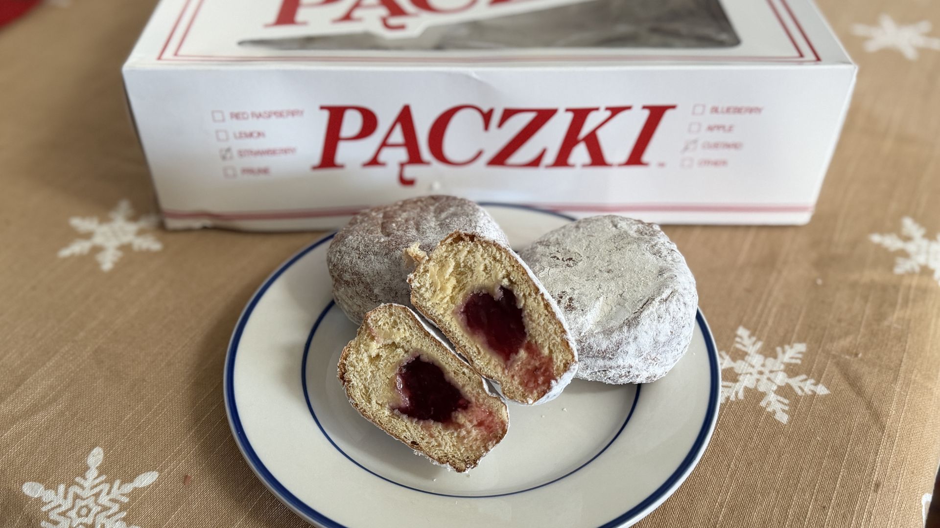 Powdered sugar-covered paczki doughnuts on a white plate with blue rim, one cut in half showing bright red strawberry jelly filling, box labeled "PACZKI" in red on table with snowflake-patterned cloth.