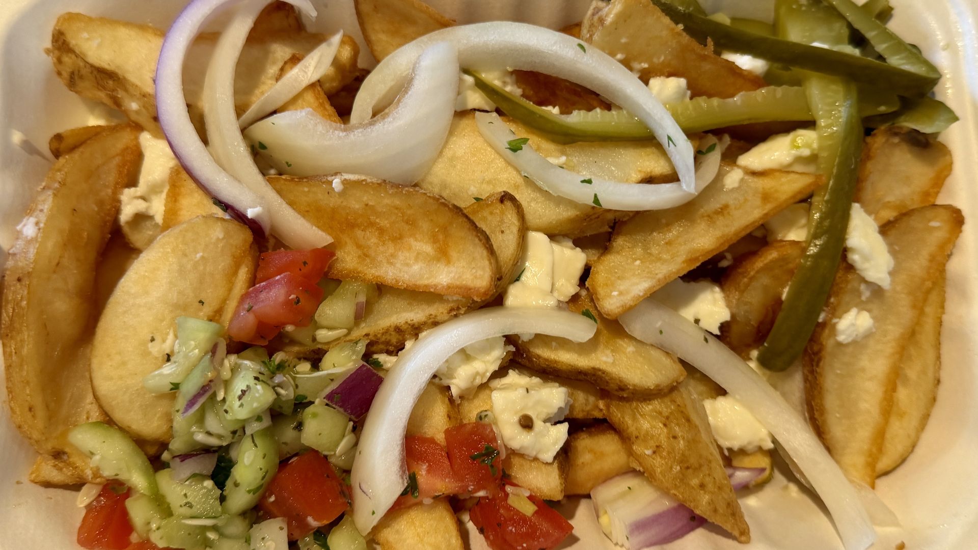 Takeout box with golden potato wedges topped with feta, large white onion rings, a tomato-cucumber salsa, and sliced pickled green peppers.