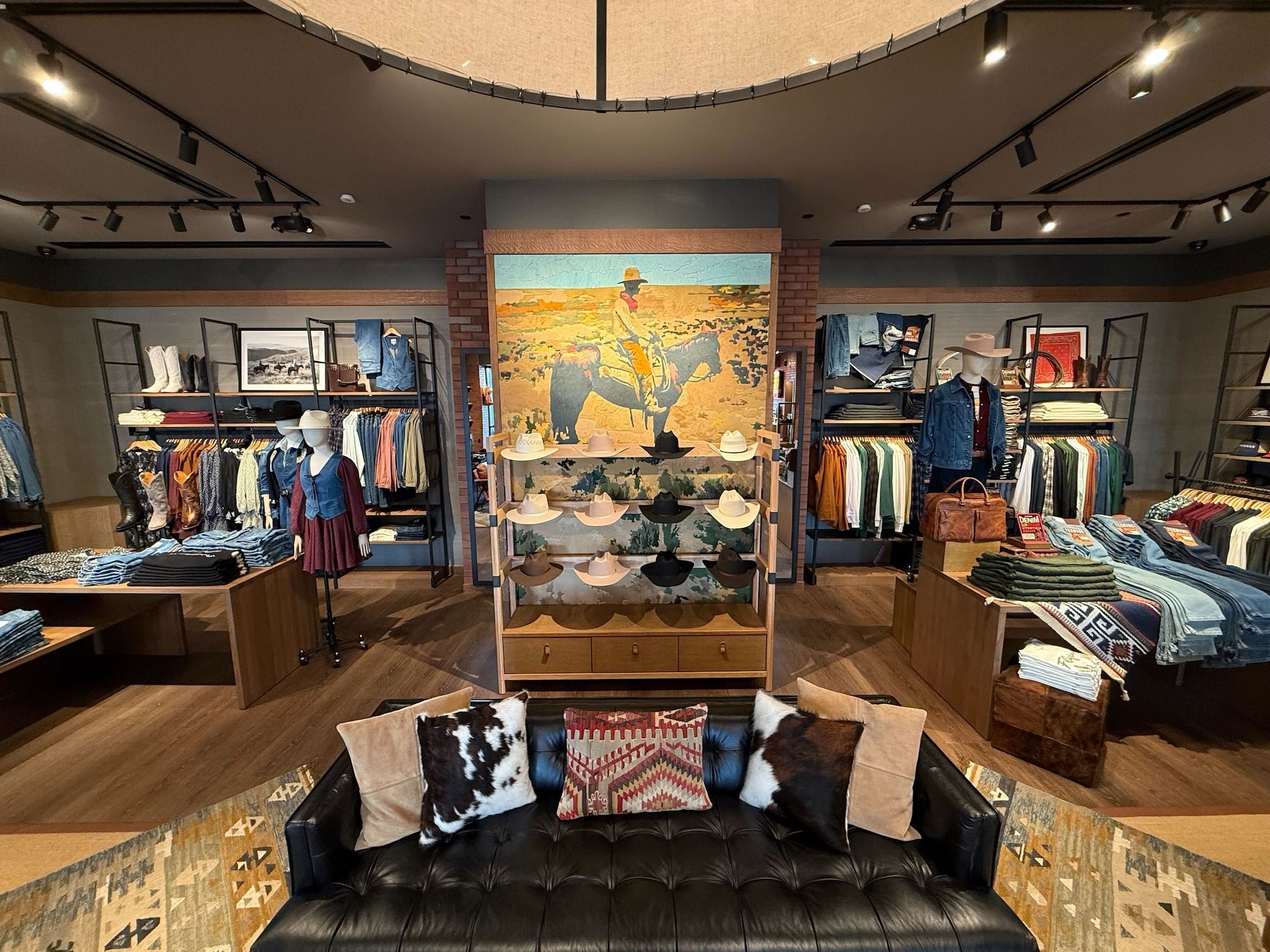 Interior of a Western-themed clothing store with cowboy hats, denim, boots, mannequins wearing Western outfits, and a leather couch with patterned pillows in the foreground.