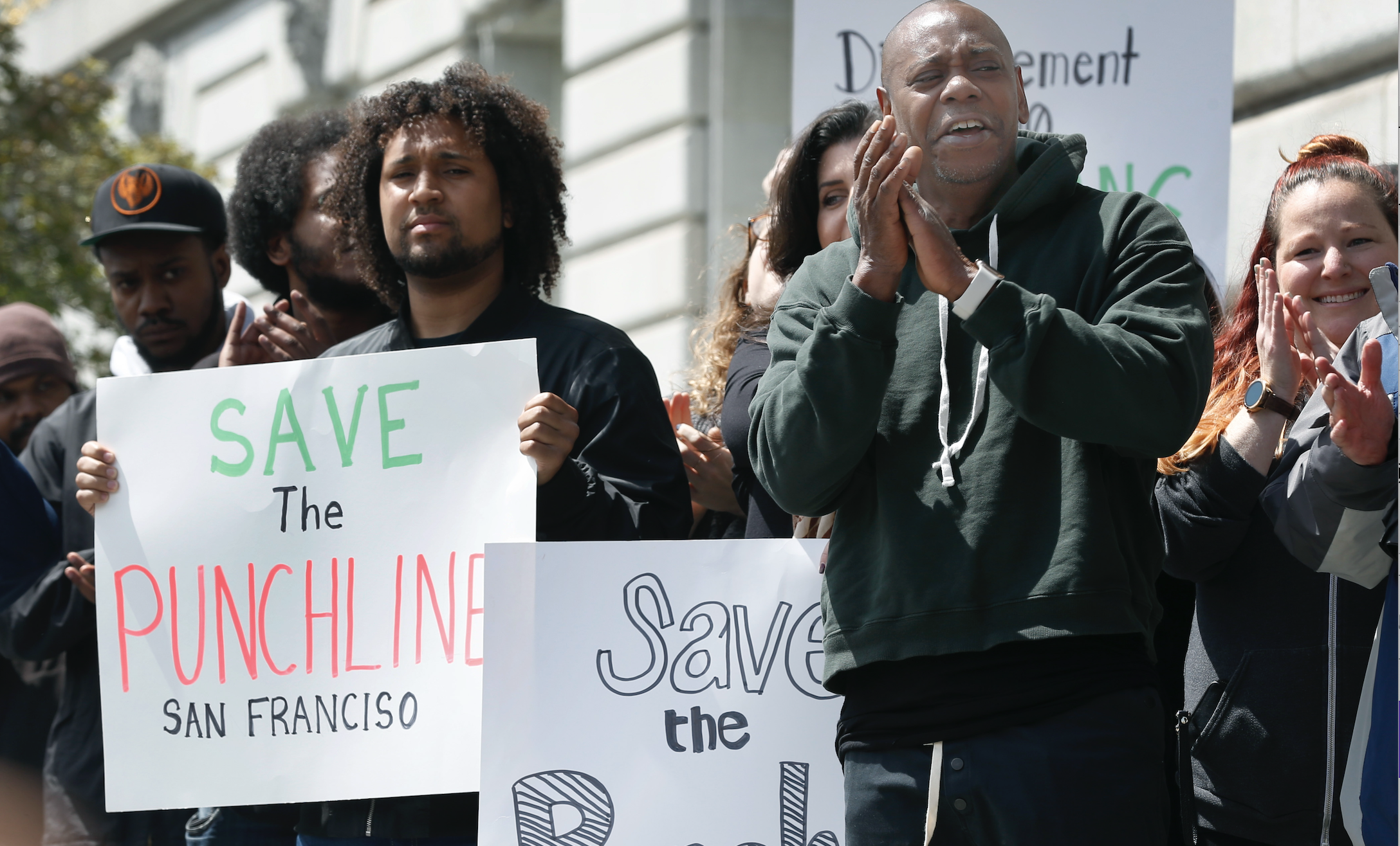 Dave Chappelle claps while standing, surrounded by people holding "Save the Punch Line" signs