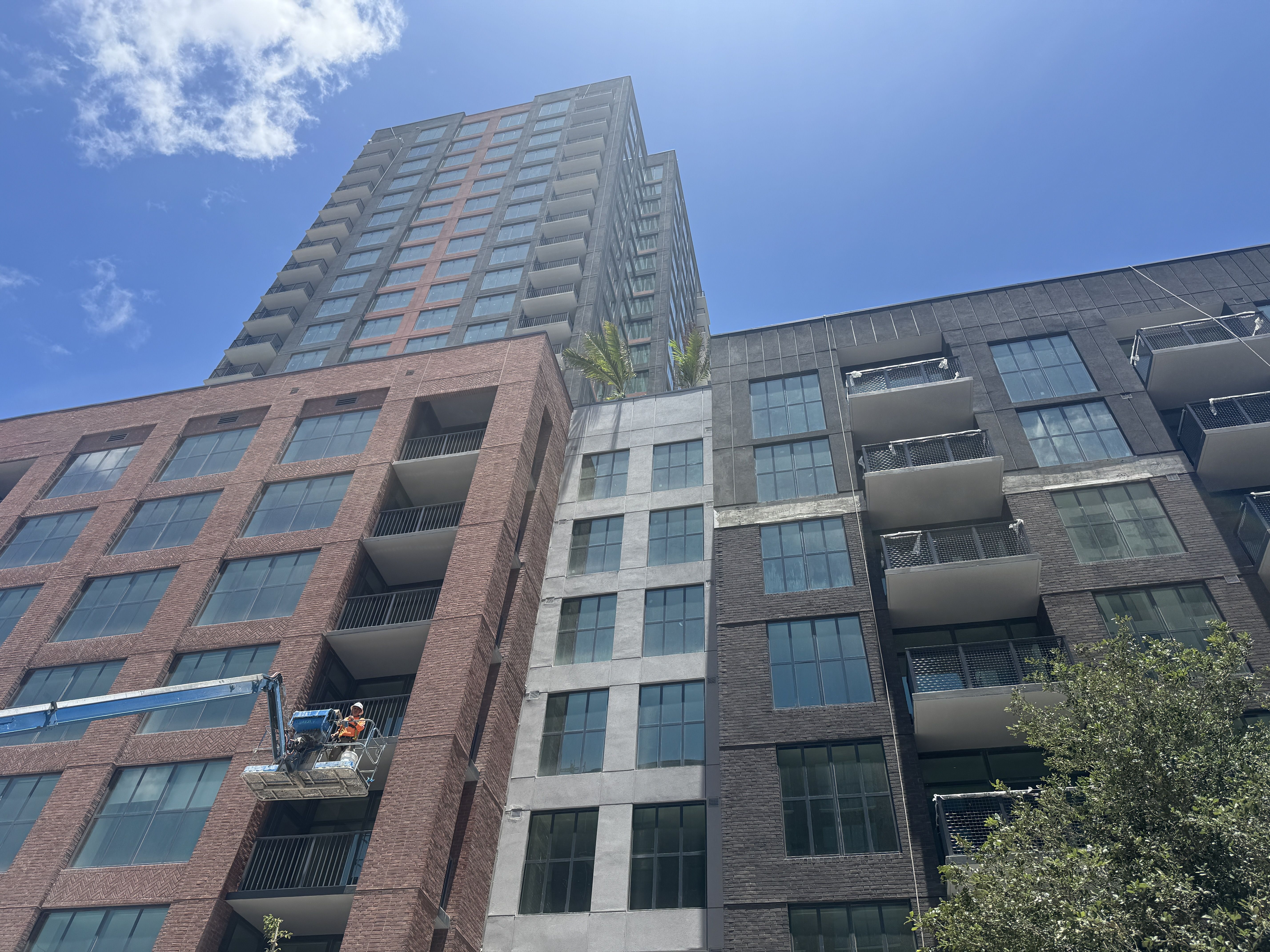 Low-angle shot of a tall apartment building with red brick and gray panels, many balconies and glass windows. Sky above; a palm tree on a terrace and workers on a construction lift at bottom left.
