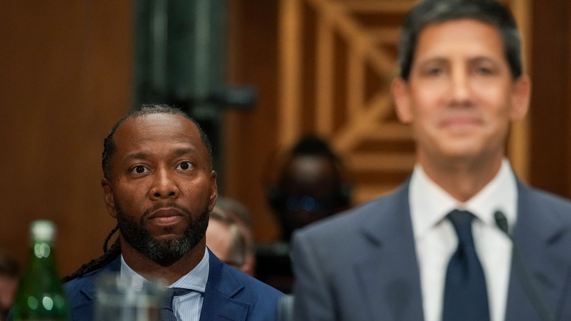 A man in a dark blue blazer over a light blue shirt with a dark tie sits in a congressional hearing room. A man wearing a gray blazer, white shirt and dark tie sits in the foreground, slightly out of focus. 