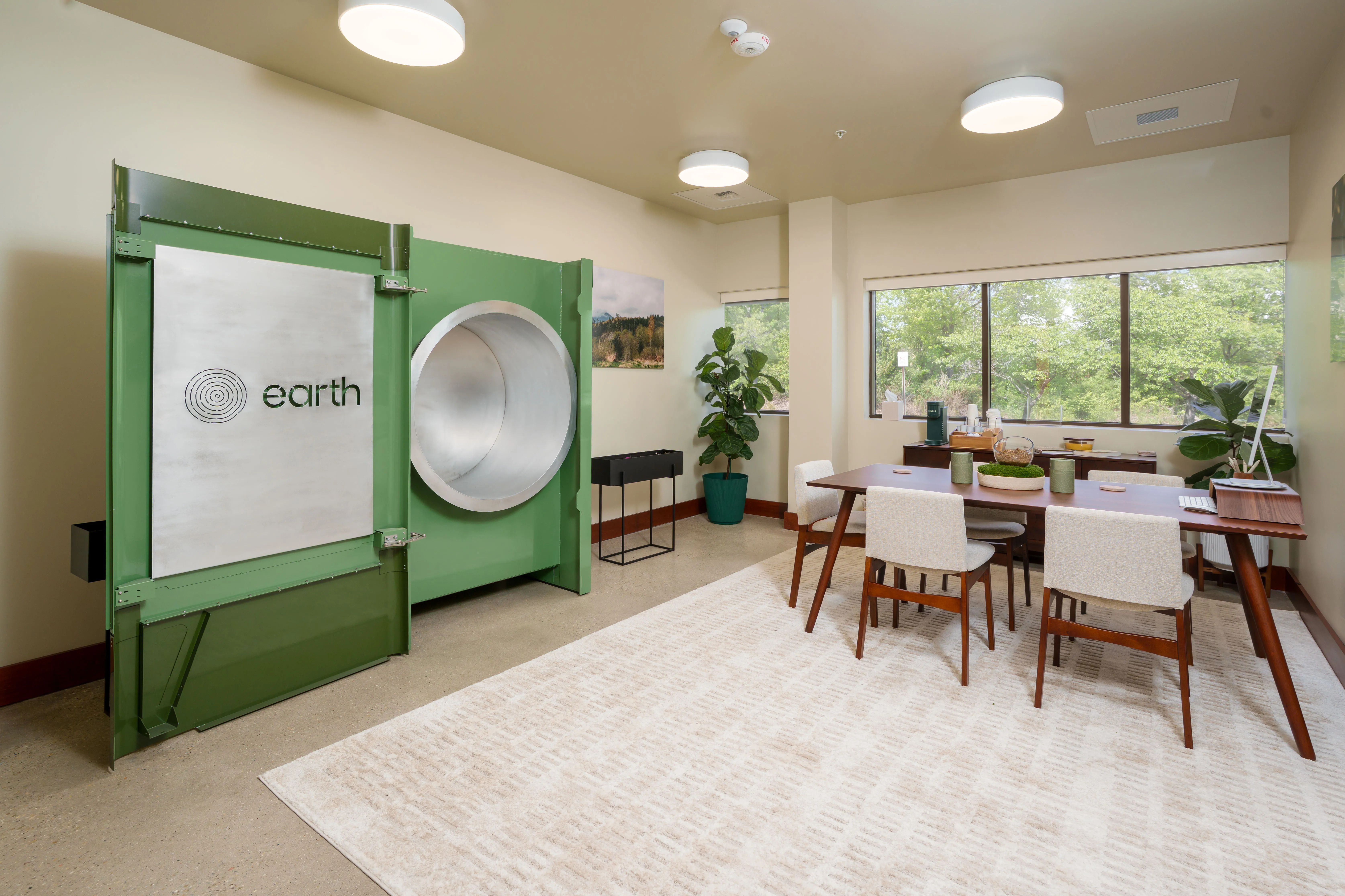 Bright room with a large green machine labeled earth and a circular metal opening; a wooden conference table with white chairs, plants, and windows showing green trees outside.