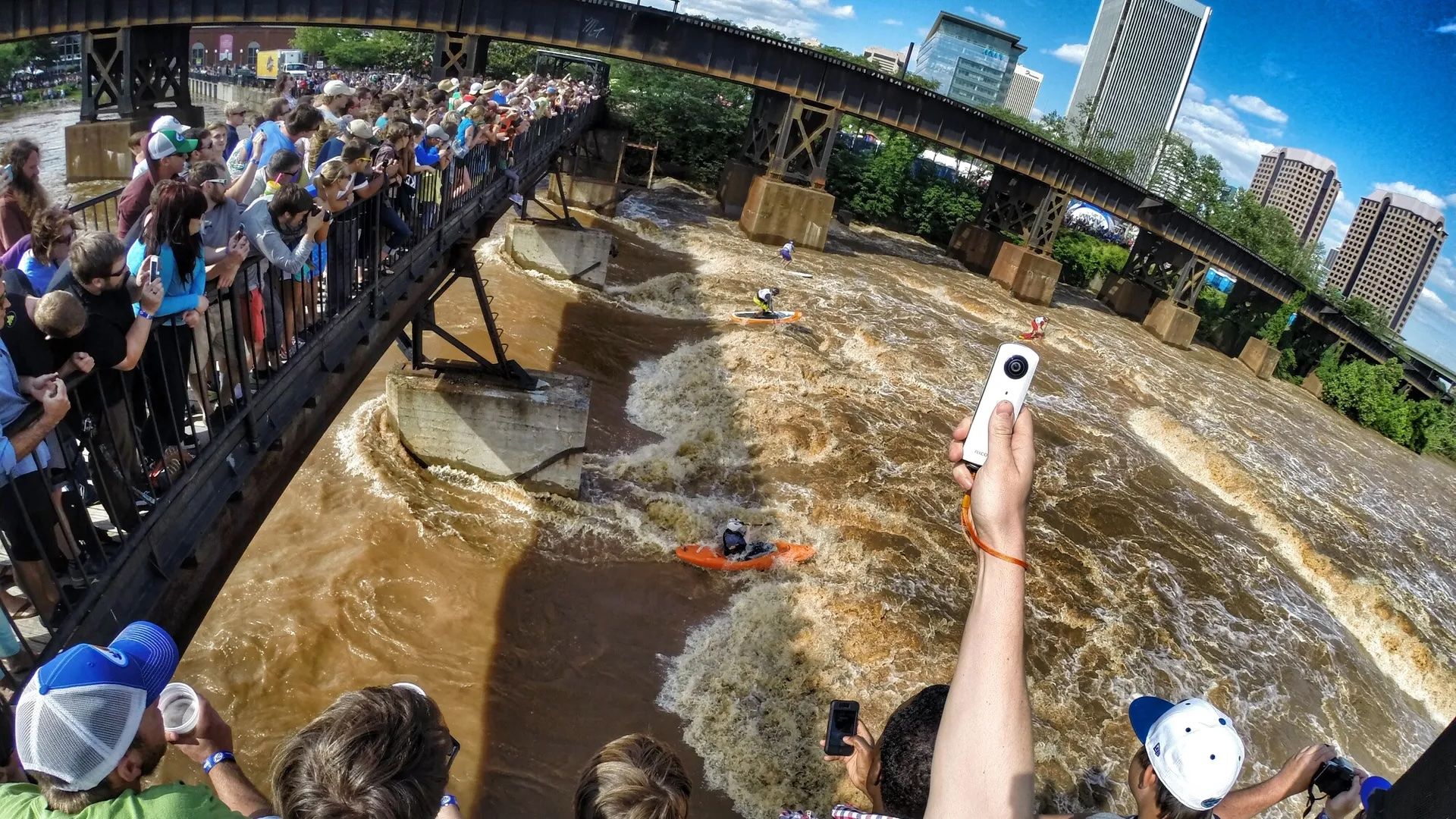 Crowded pedestrian bridge over a muddy, fast river; orange kayaks ride rapids beneath as onlookers lean over railings; a person on the right holds a white action cam toward the scene.