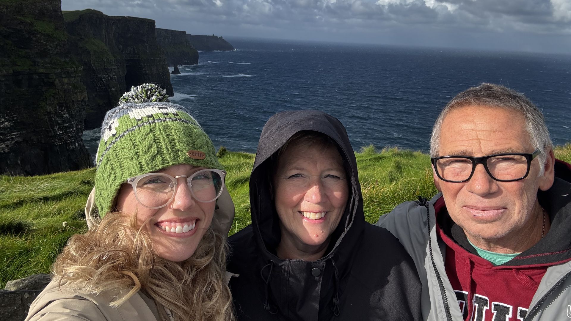 Three adults smiling for a selfie near tall green cliffs by a deep blue sea under a partly cloudy sky, wearing jackets and a green knit hat.