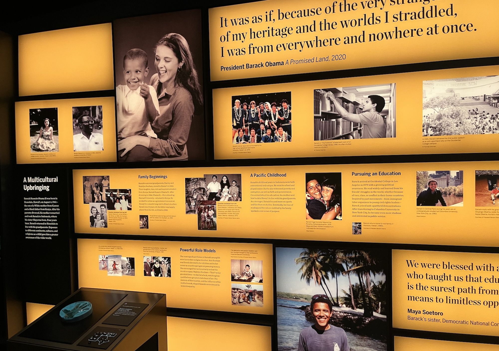 Exhibit wall with bright yellow panels showing black-and-white family photos and a large quote about heritage; smaller captions adorn the display, while a black console with a blue round object sits in front.