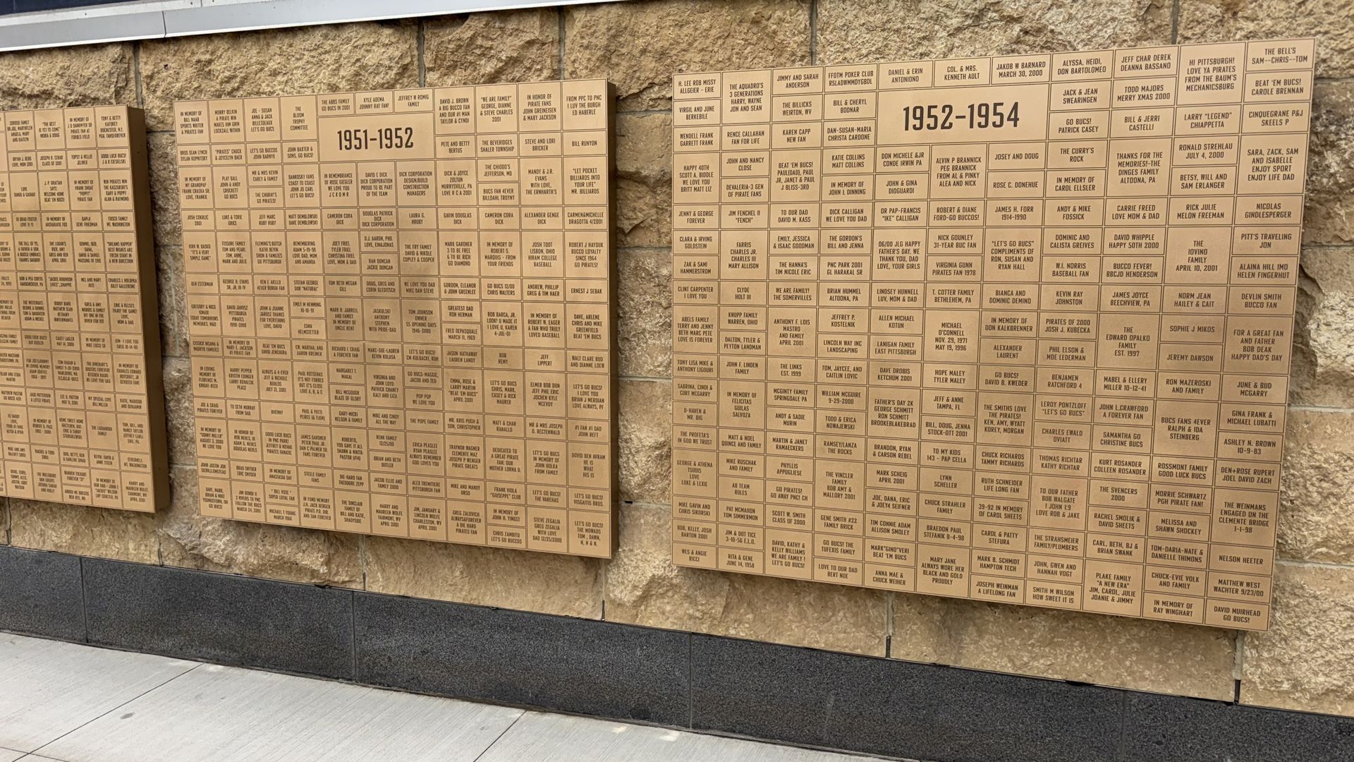 Beige stone wall with large bronze plaques arranged in columns, listing names under year headings such as "1951-1952" and "1952-1954" in small print.