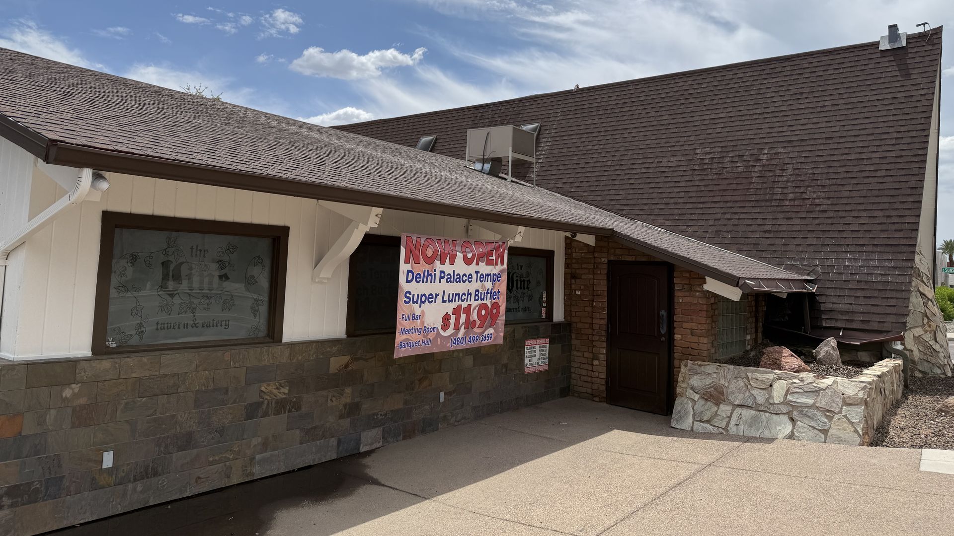 A single-story storefront with a brown shingled roof, beige siding, and a large 'NOW OPEN' banner for Delhi Palace Tempe, with an A-frame section viewed from the side; blue sky with scattered white clouds overhead.