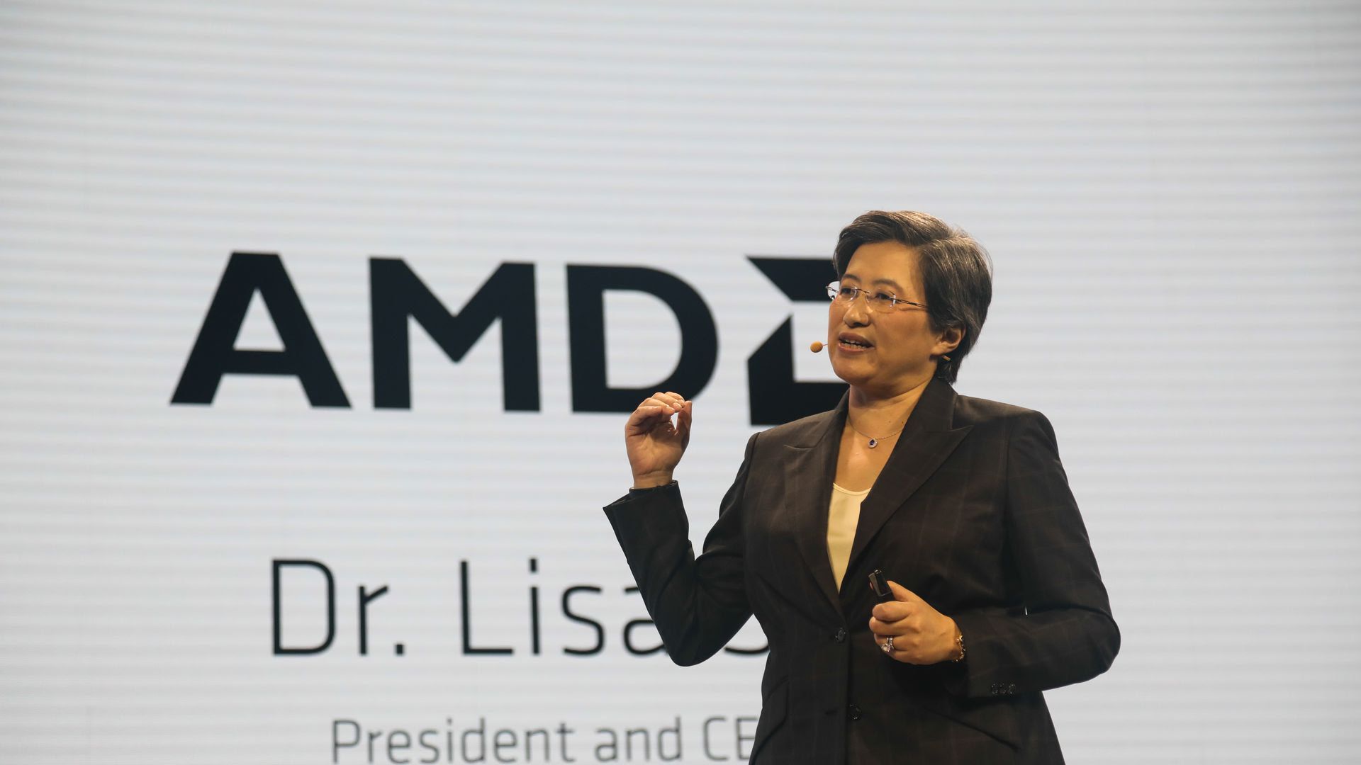 This image shows Su speaking on stage while wearing a blazer and microphone. The large screen behind her shows the AMD logo and her name.