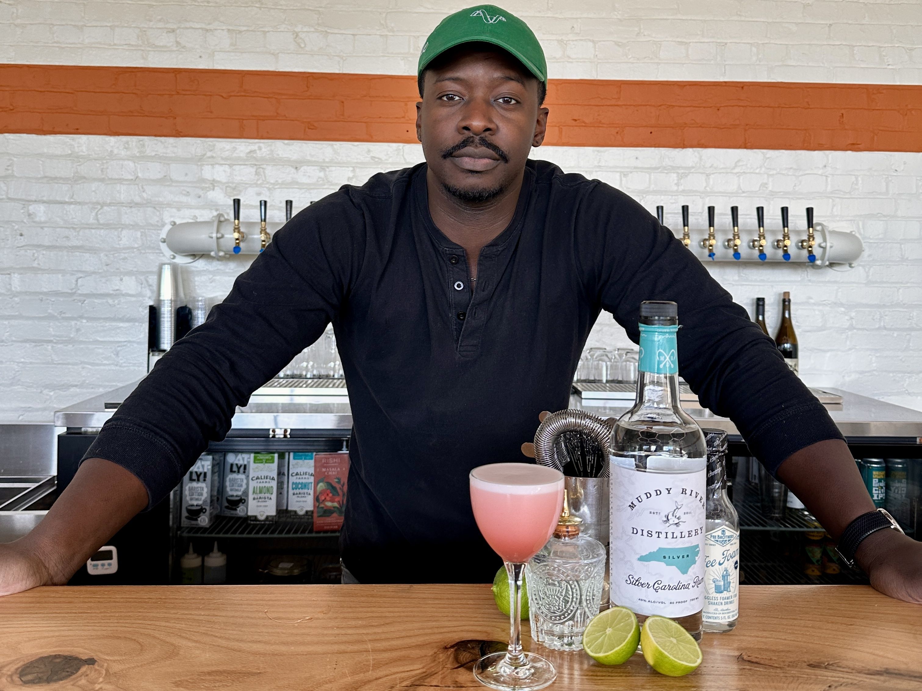 Man with a green cap sits behind a blue counter, arms resting on the surface, in an industrial cafe with exposed pipes, hanging lights, a white brick wall with an orange stripe, and bar equipment. Lorem Ipsum's founder opens new and/or cafe at Innovation Barn.