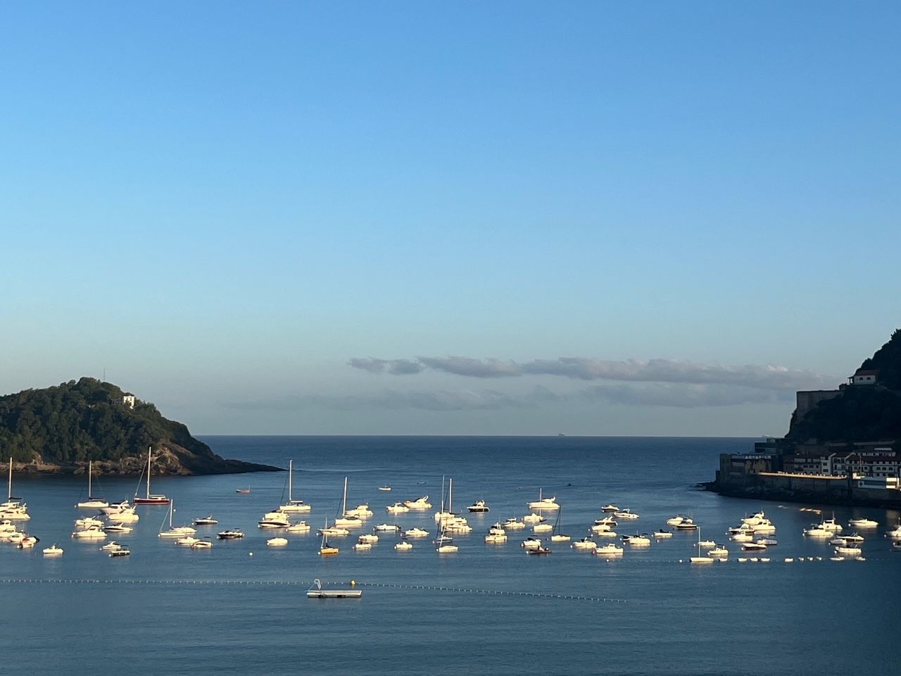 Calm blue bay dotted with numerous white boats anchored; tree-covered headlands frame the left and right, with a coastal town along the right shore and a clear sky above.