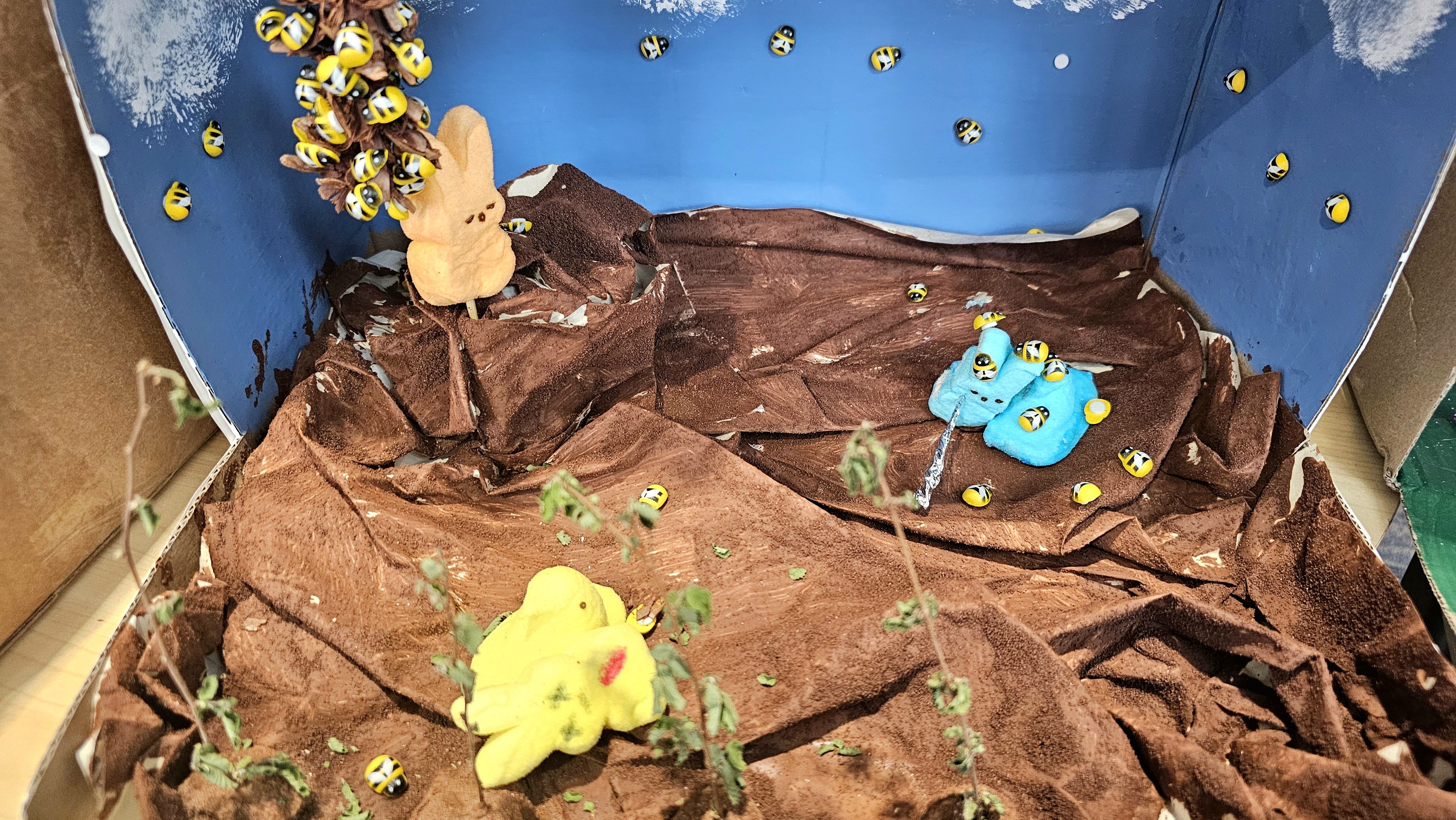 Small diorama inside a blue box with brown faux rock terrain, many yellow-and-black bees, a beige plush bunny on the left, two blue plush figures on the right, and a yellow plush at the front.