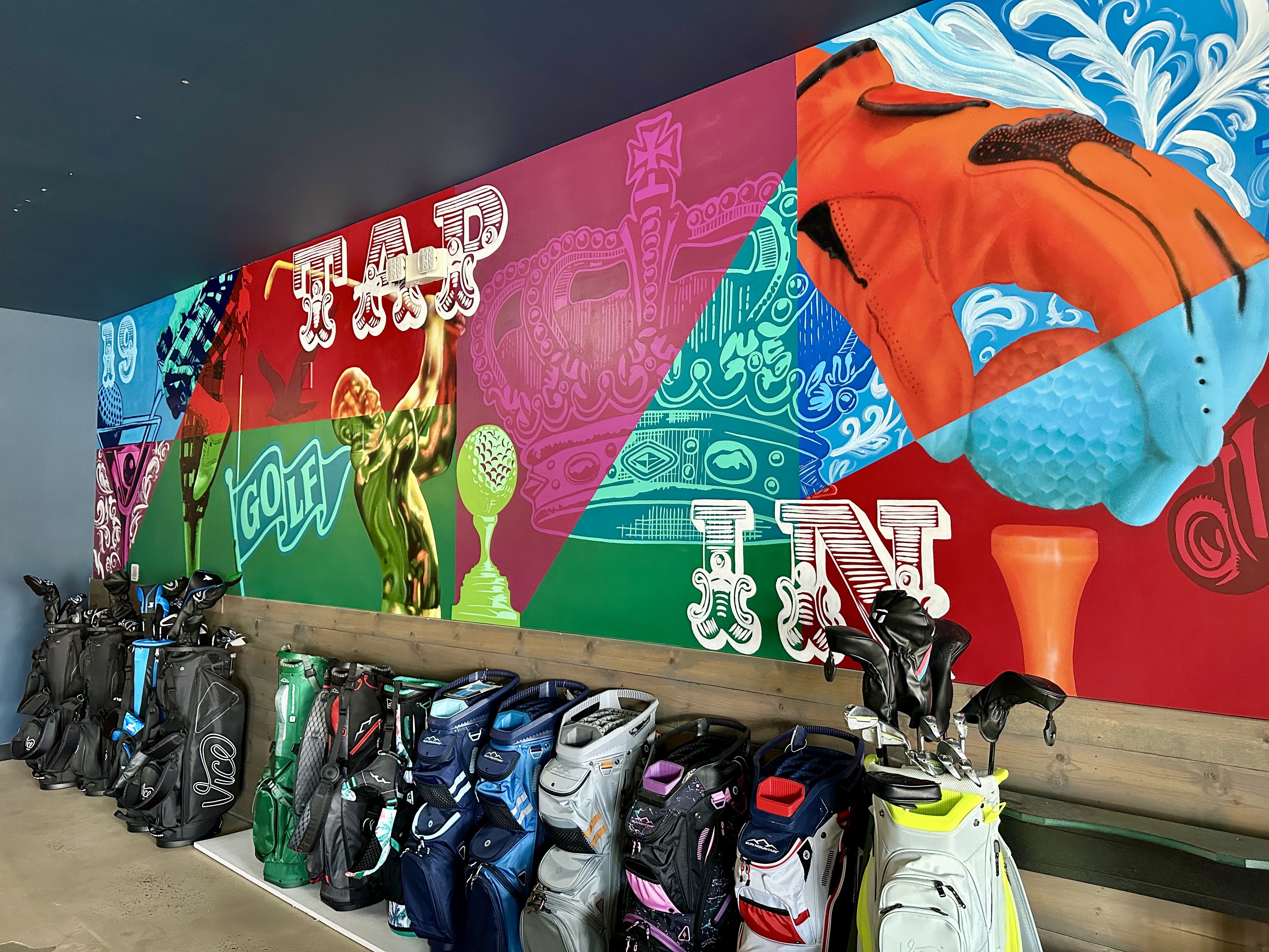 Colorful indoor golf club storage with multiple golf bags and clubs lined up against a wall featuring vibrant mural art with golf imagery and text including "TAR" and "IN".