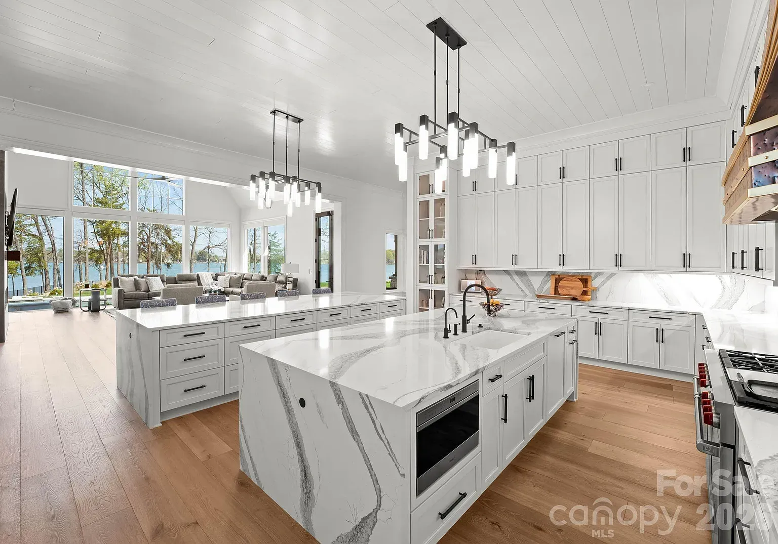 Bright kitchen with white cabinets, marble countertops, and a large island with a sink and built-in microwave. Black hardware, light wood floors, two modern chandeliers, and expansive windows framing a lake view.