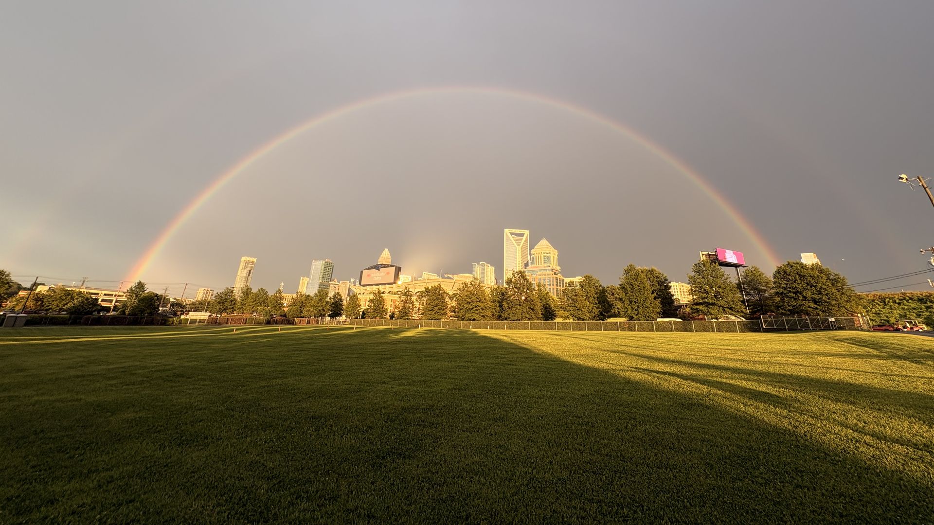 Wide shot of a city skyline at sunset with a clear double rainbow arching across a pastel sky; a grassy field in the foreground and trees line the bottom.