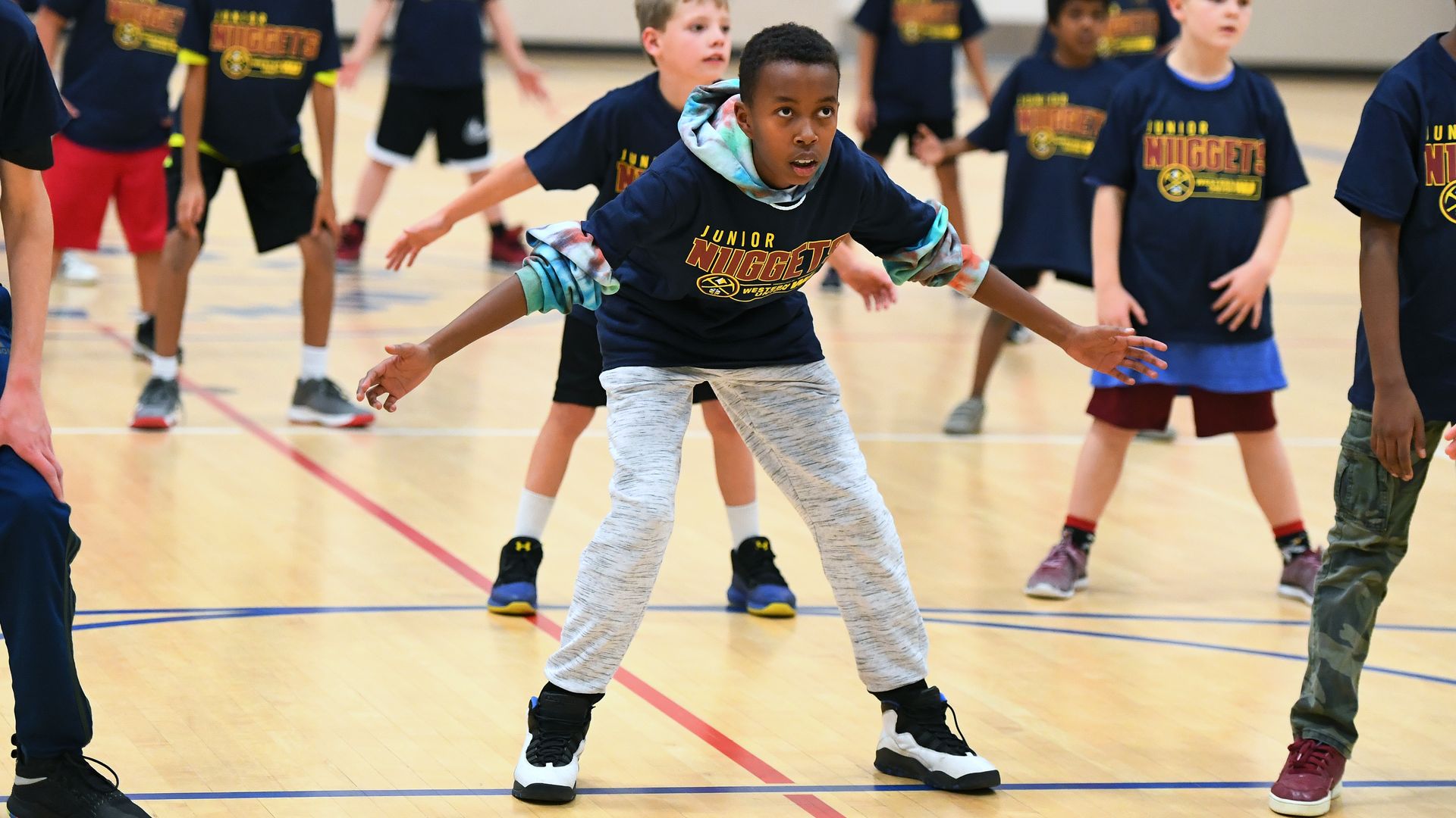 A child in a navy shirt and gray sweatpants bends down as if stretching inside a gym, surrounded by several other children in similar clothing.