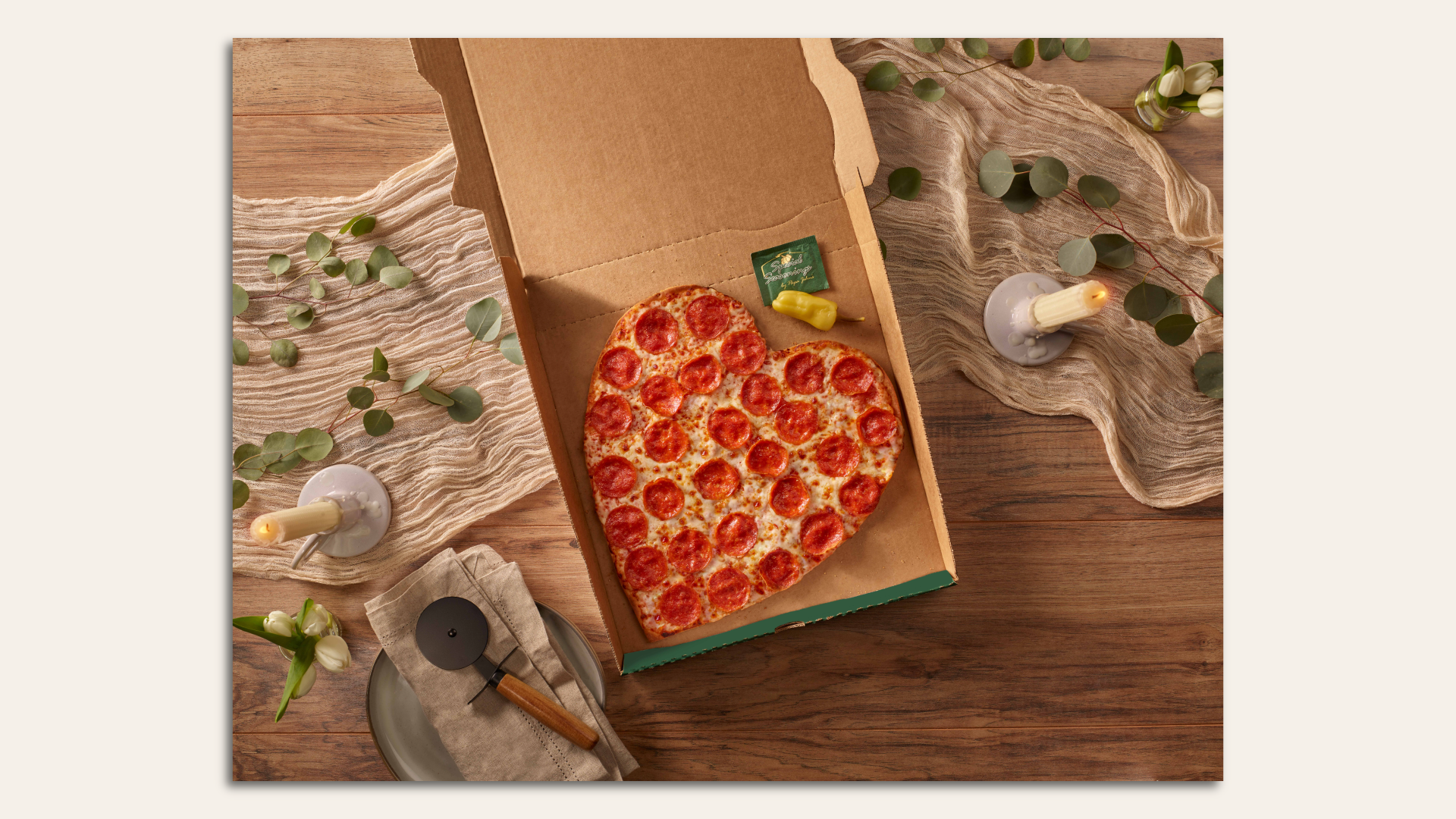 A heart-shaped pizza in a pizza box surrounded by the trappings of a candlelight dinner.