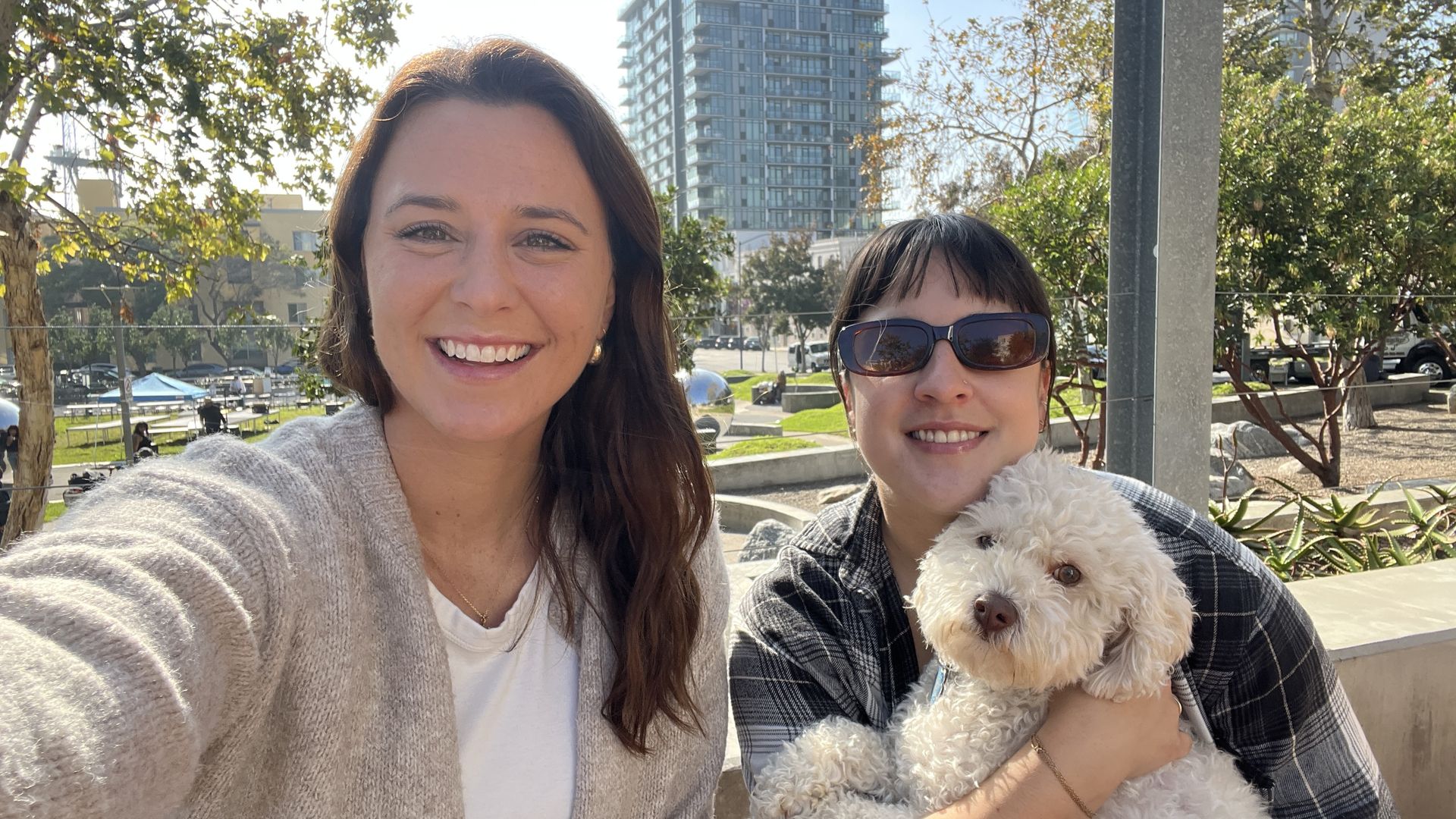 Two smiling women outside on a sunny day; one wears a beige sweater, the other sunglasses and a plaid shirt, holding a fluffy white dog. Trees and buildings in the background.