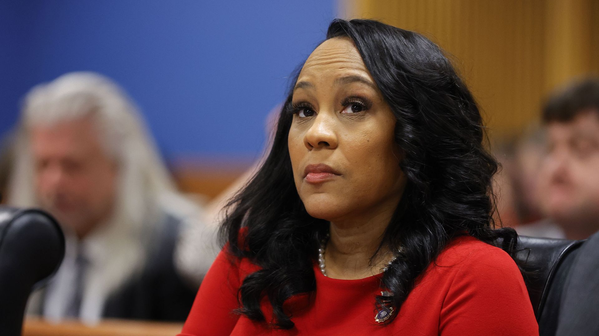 A woman wearing a red dress and pearls sits in a courtroom and looks toward the camera