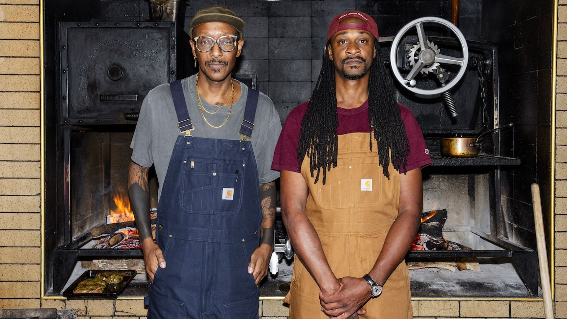Two men stand in front of a large grill with a fire burning behind them. Both wear overalls—navy blue and tan—with t-shirts and caps in a kitchen setting.