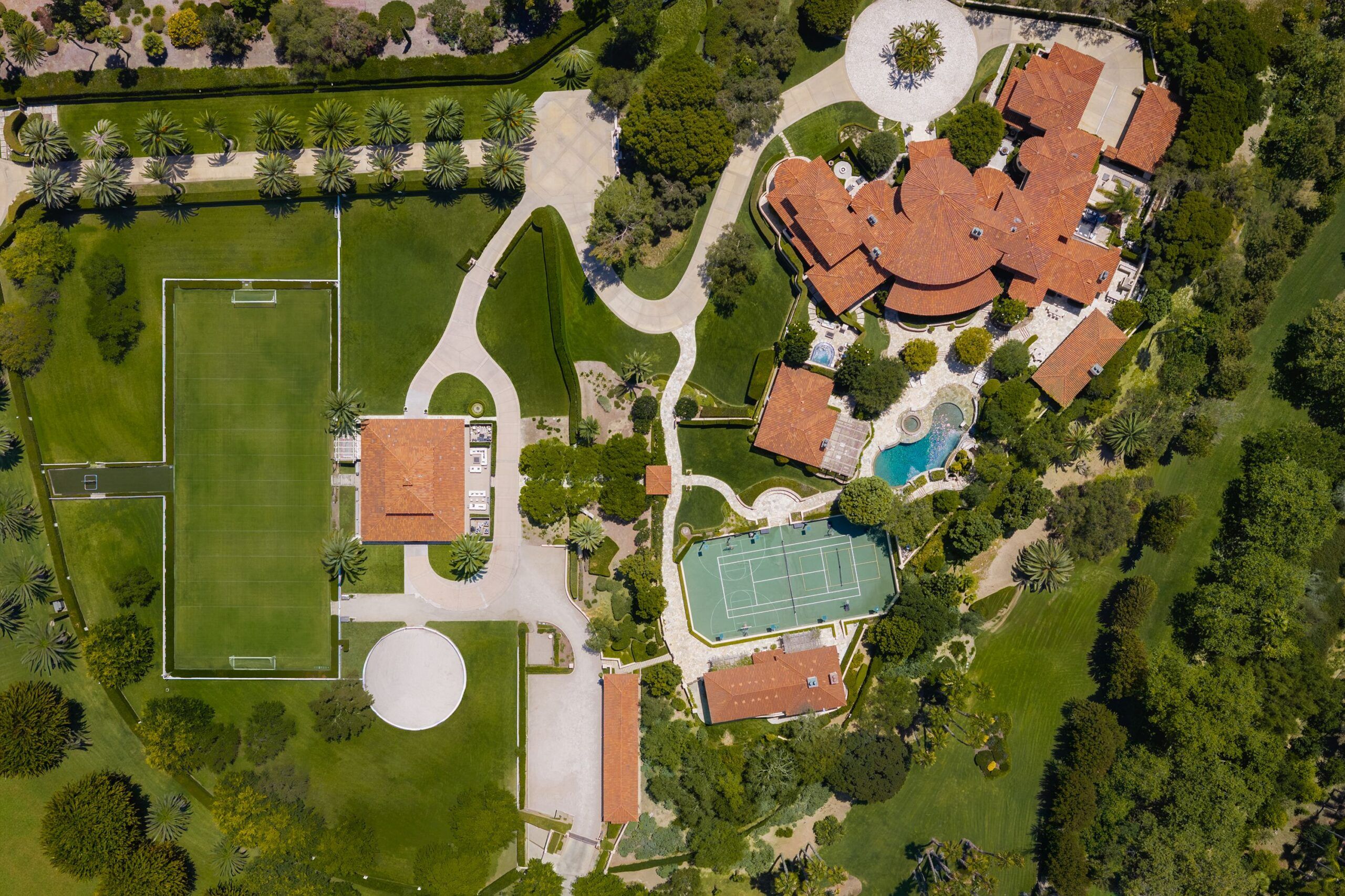 Aerial view of a luxury estate with red-tiled mansion, curved circular driveway, palm-lined walkways, expansive manicured lawns, a swimming pool, tennis courts, and a large sports field.