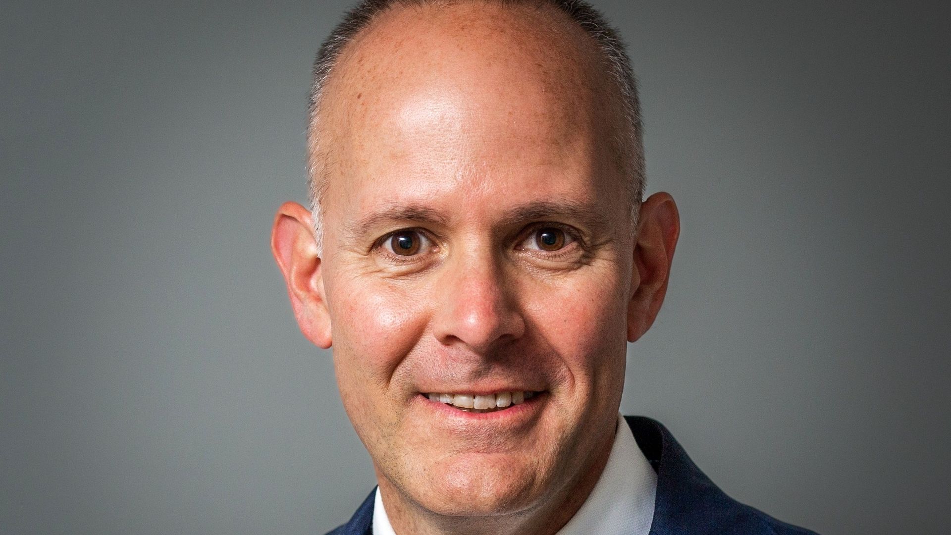 Close-up of a smiling man in a dark blue suit and a striped red, white, and blue tie, standing against a neutral gray background.