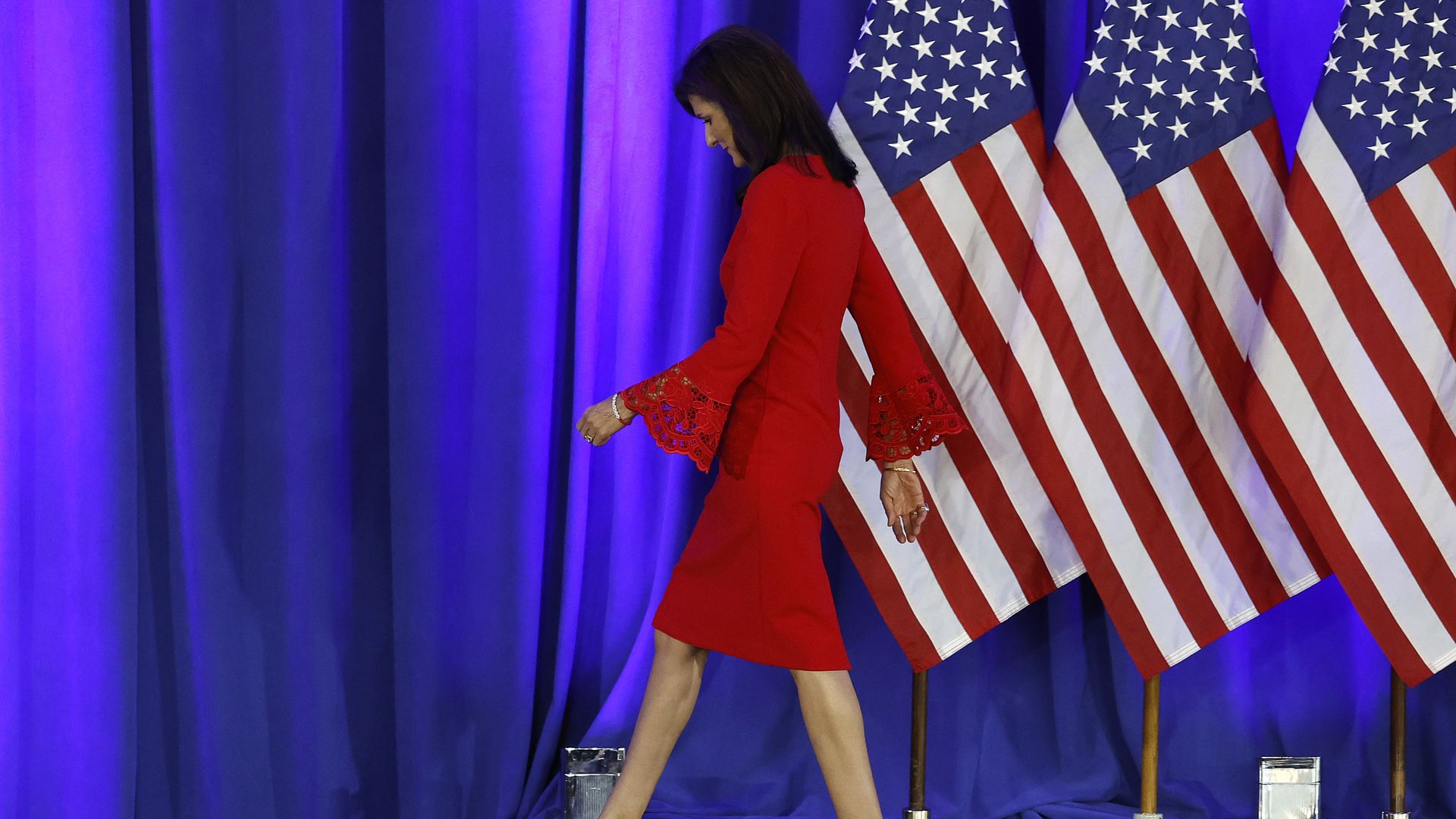 Nikki Haley, wearing a red dress with American flags in the background, walks off of a stage.