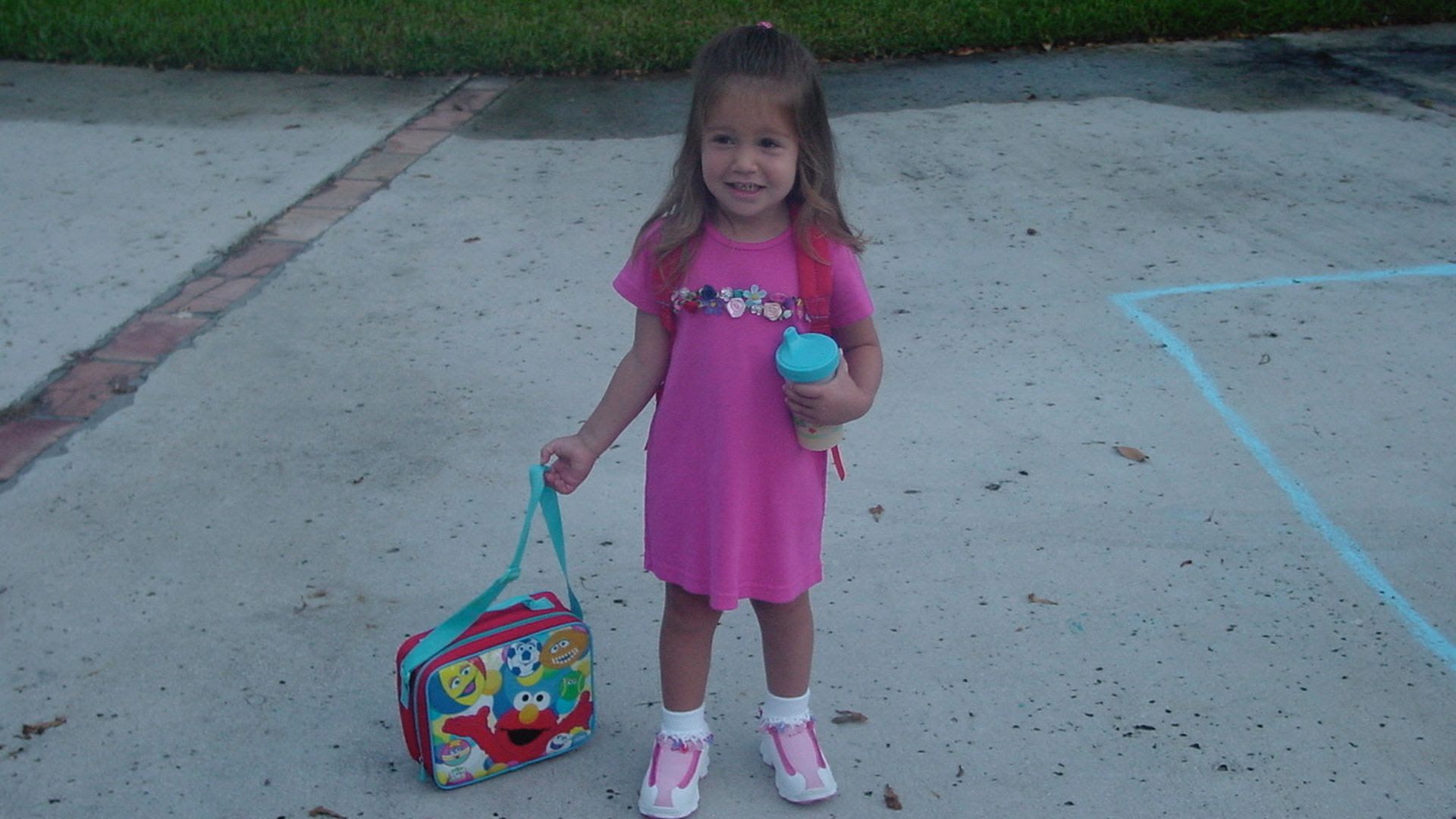 Smiling little girl in a pink dress holds a teal water bottle and bag, standing on a concrete driveway with a colorful lunchbox at her feet; a blue chalk line is on the pavement behind her.