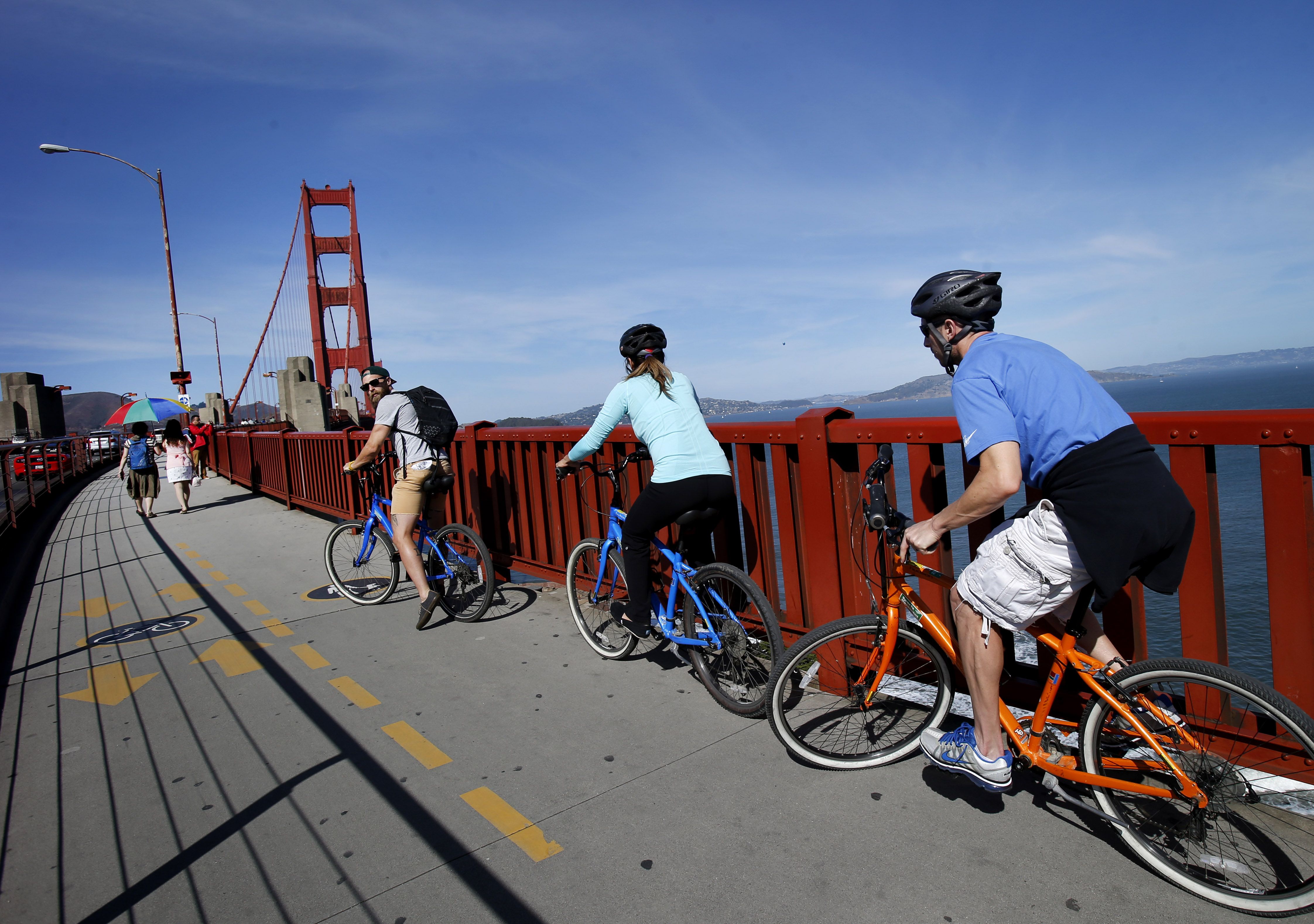 Three cyclists, two on blue bikes and one on an orange bike, ride across the Golden Gate Bridge on a sunny day, with pedestrians walking nearby and blue sky above.