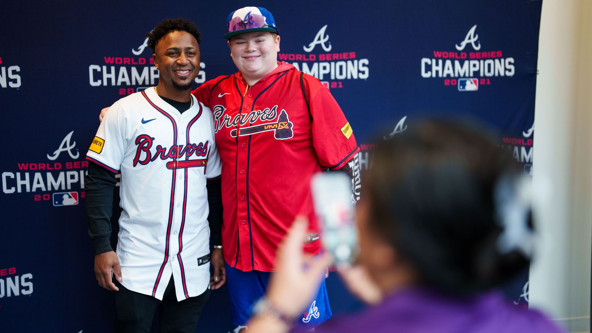 Two men smiling and posing in Atlanta Braves jerseys, one white and one red, in front of a dark backdrop with "World Series Champions 2021" and MLB logos, while a person takes their photo.