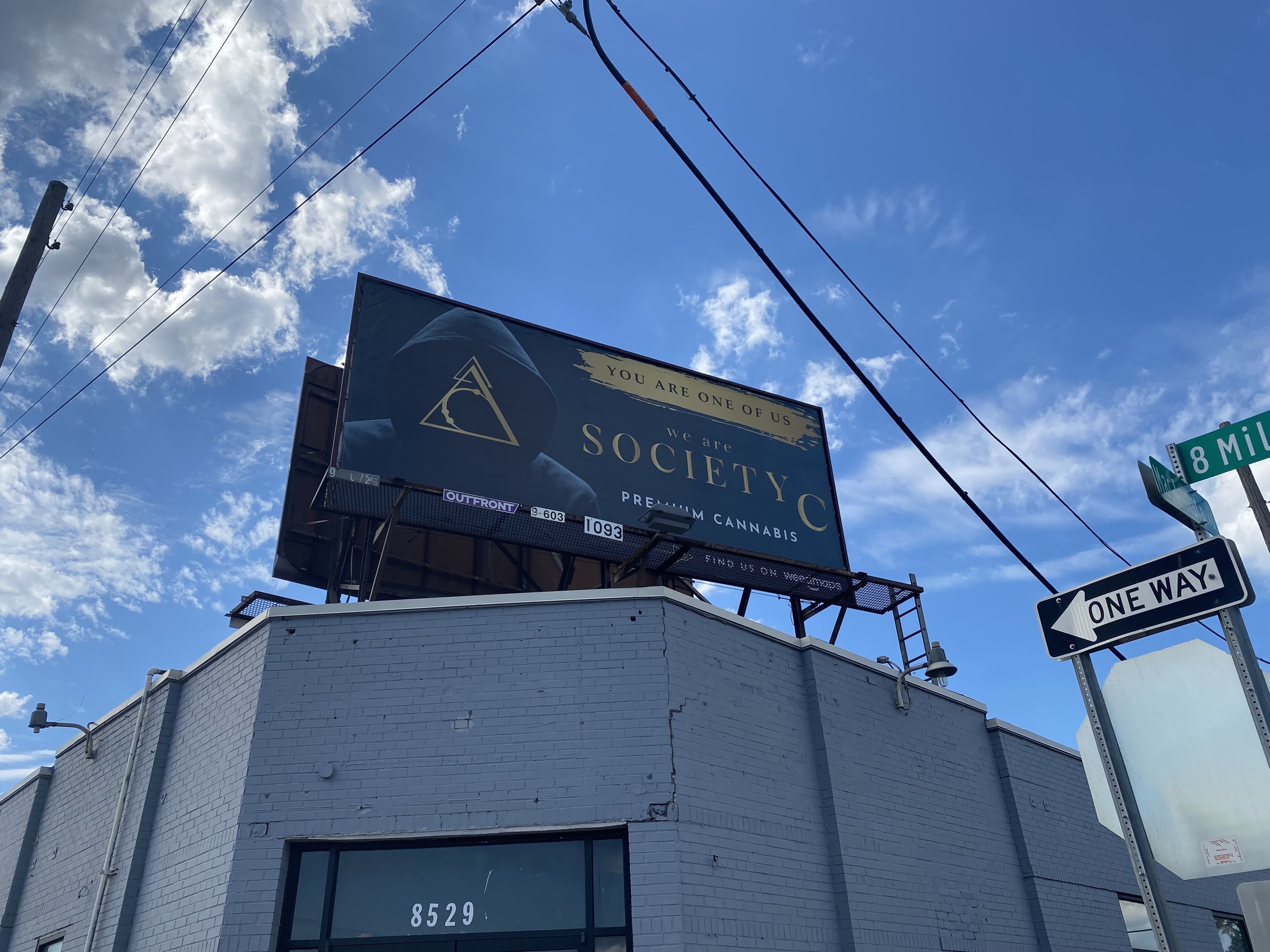 A billboard shows a hooded figure with a symbol overlaid and "We are Society C" cannabis 