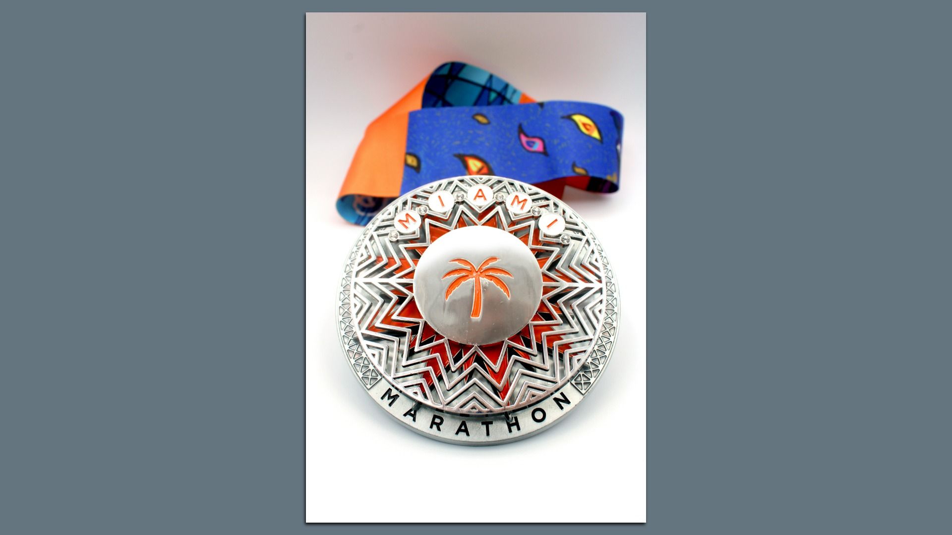 Silver Miami Marathon medal with orange palm tree in center, geometric sunburst pattern, and multi-colored blue, orange, and red ribbon on white background.
