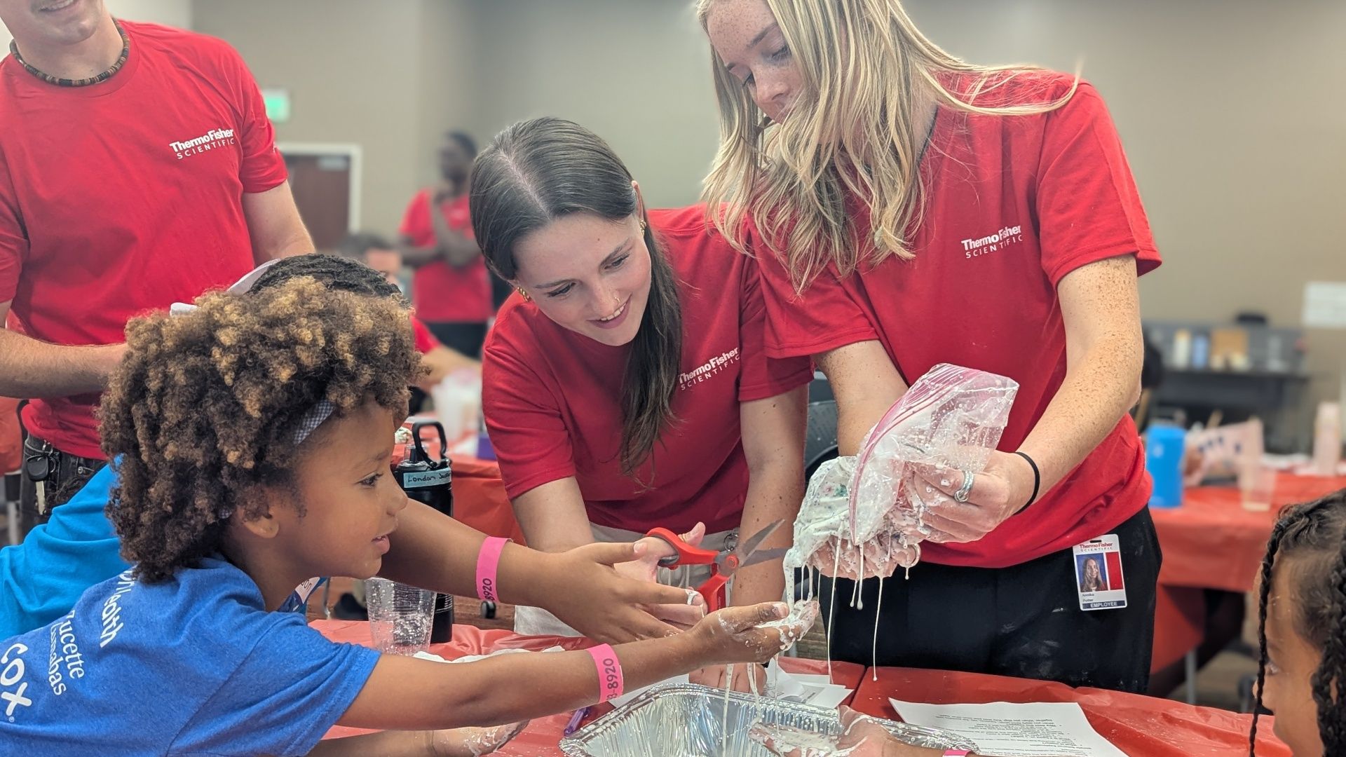 Children and volunteers in red ThermoFisher shirts participate in a hands-on science activity, cutting with scissors and handling a slime-like mixture at a table.