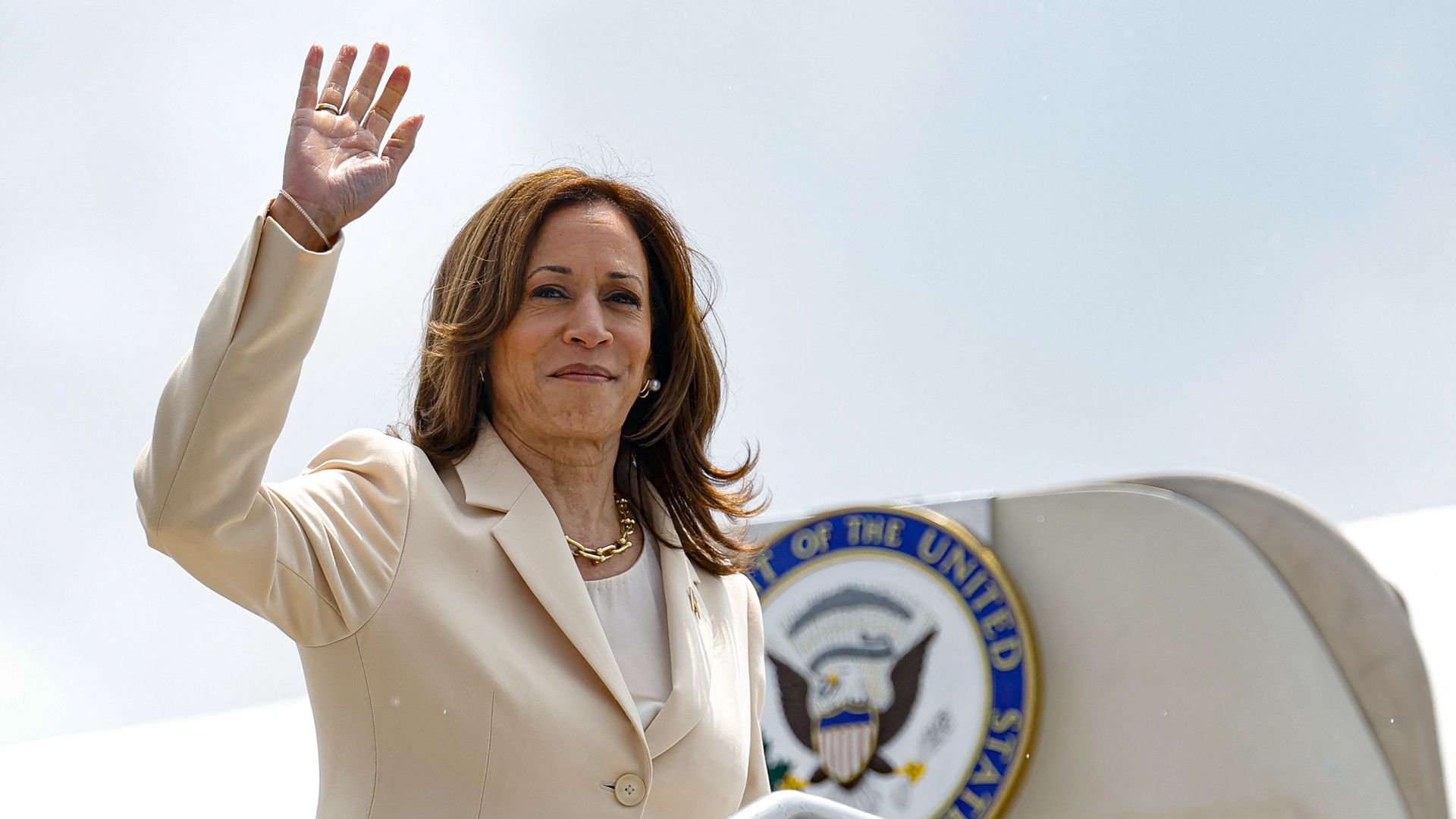 Kamala Harris, wearing a white suit and waving her hand while stepping onto an airplane with the vice presidential seal.