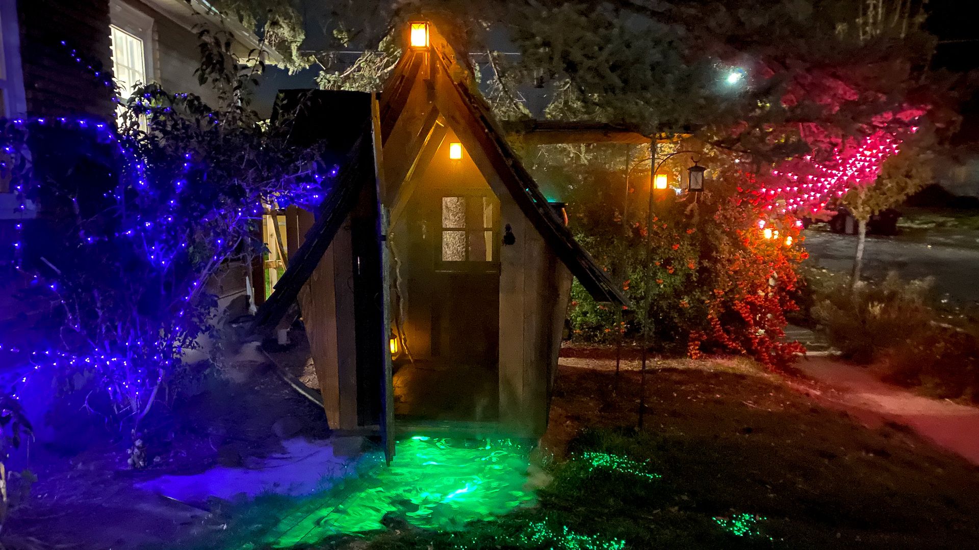 A small wooden cottage stands next to a house, with Halloween lights around it. 
