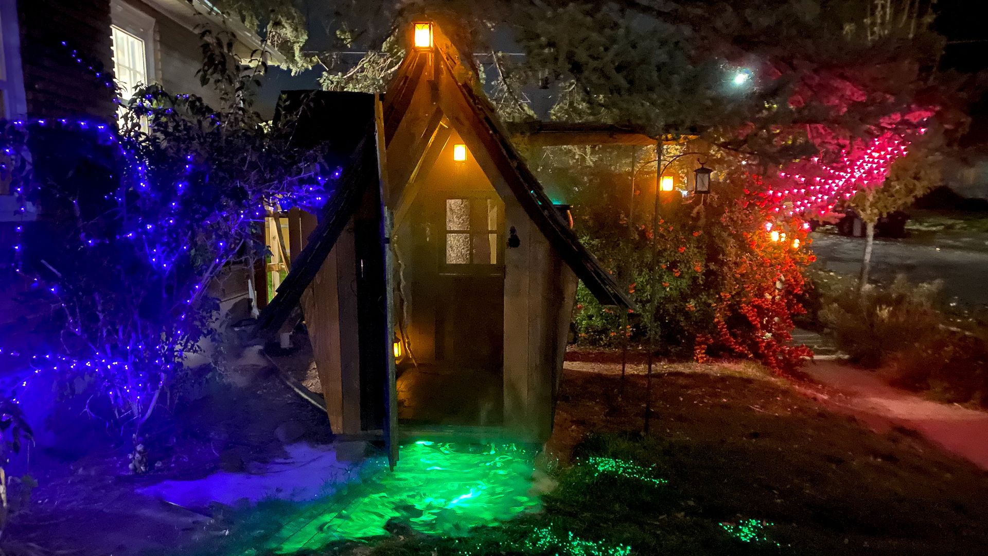 A small wooden cottage stands next to a house, with Halloween lights around it.