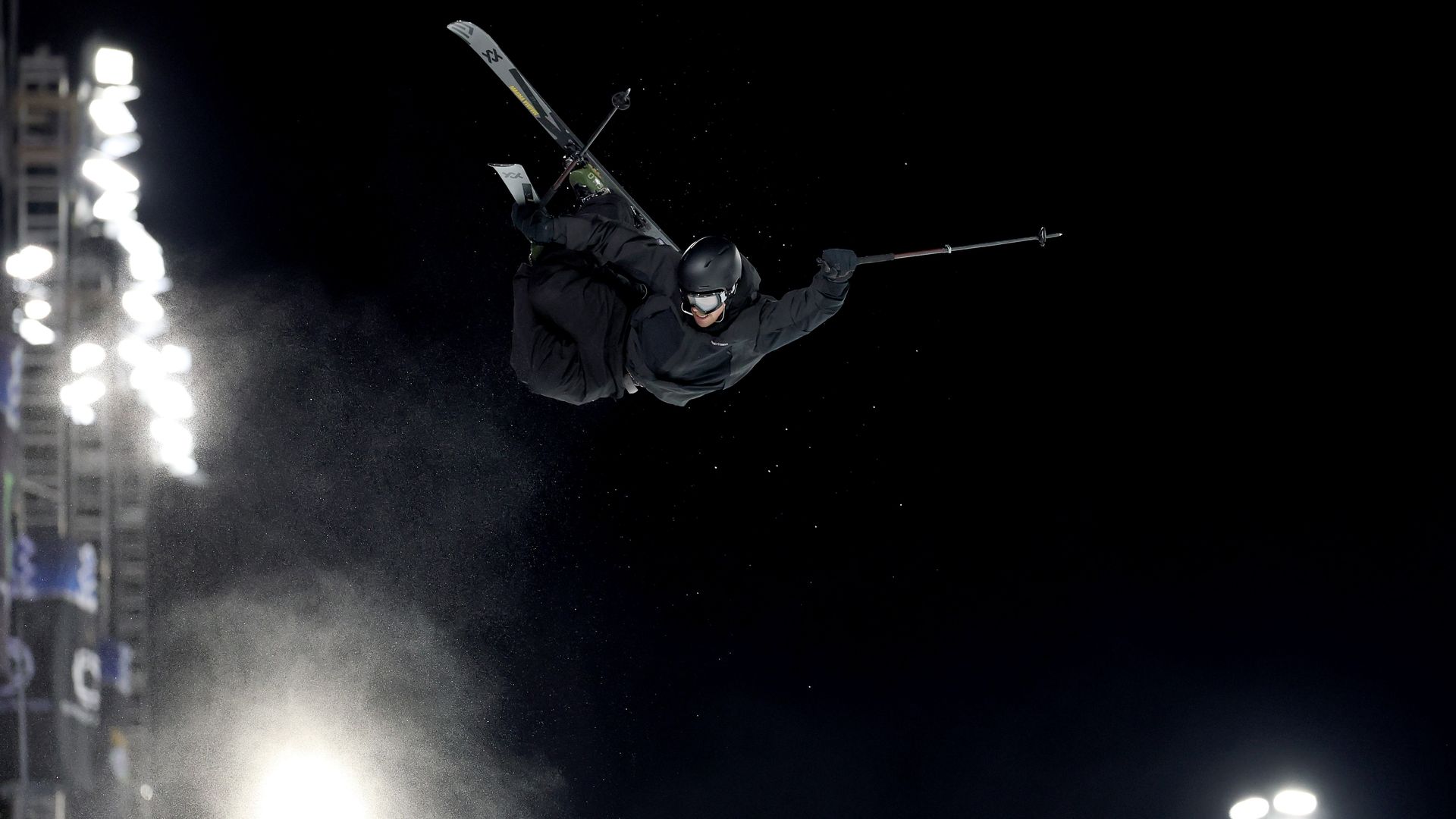 Skier dressed in black gear performs a mid-air trick at night with bright stadium lights illuminating snow particles against a dark sky.