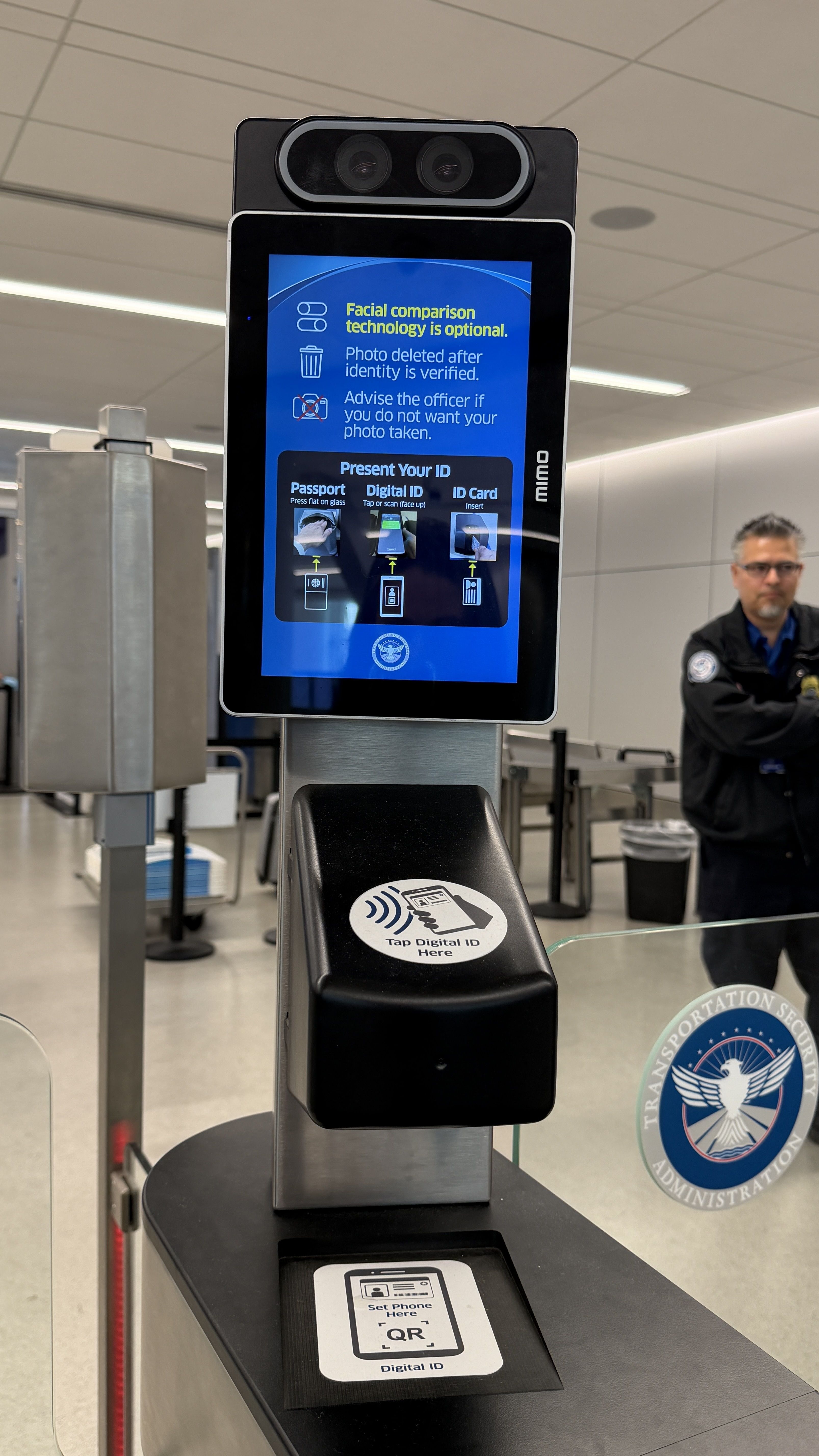Airport security kiosk with a tall black stand and blue display. Screen shows "Facial comparison technology is optional" and ID steps. Two cameras sit above; a card/QR reader is at the base.