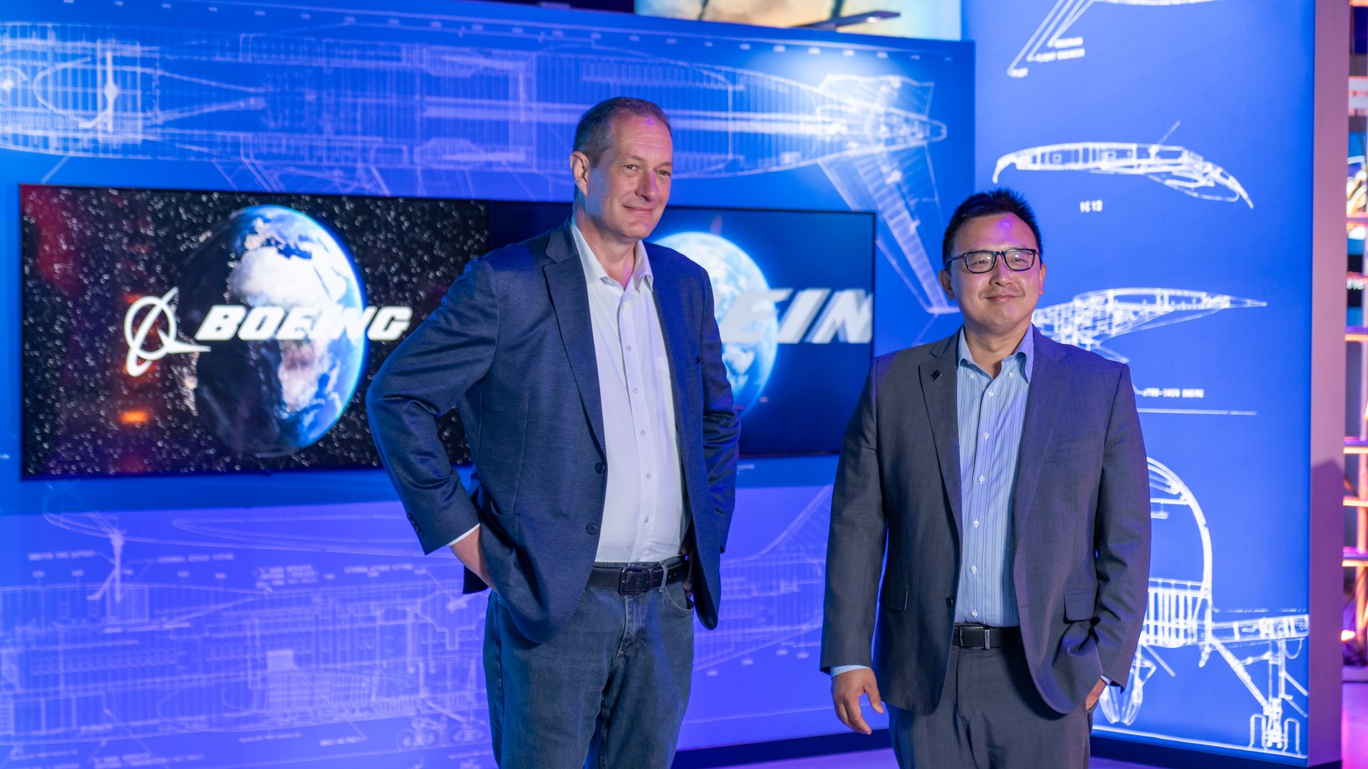 Two men in suits pose at a blue blueprint-themed exhibit. A wall of diagrams and a screen show Earth and the Boeing logo, suggesting a futuristic aerospace display.