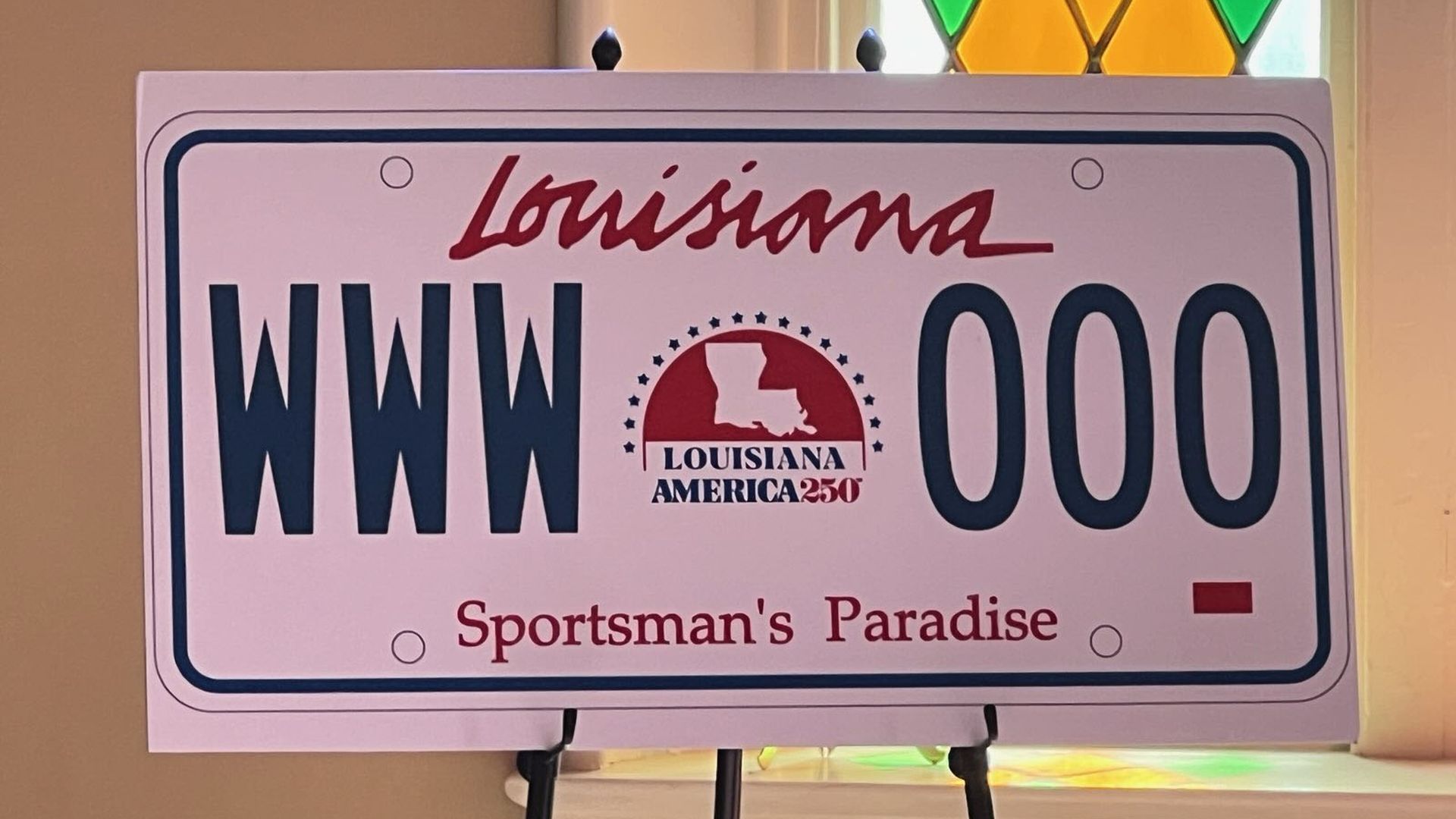 A photo of a license plate rendering on an easel. It's Louisiana license plate No. WWW 000 and has a logo at the center that says Louisiana: America 250 with an outline of the state.