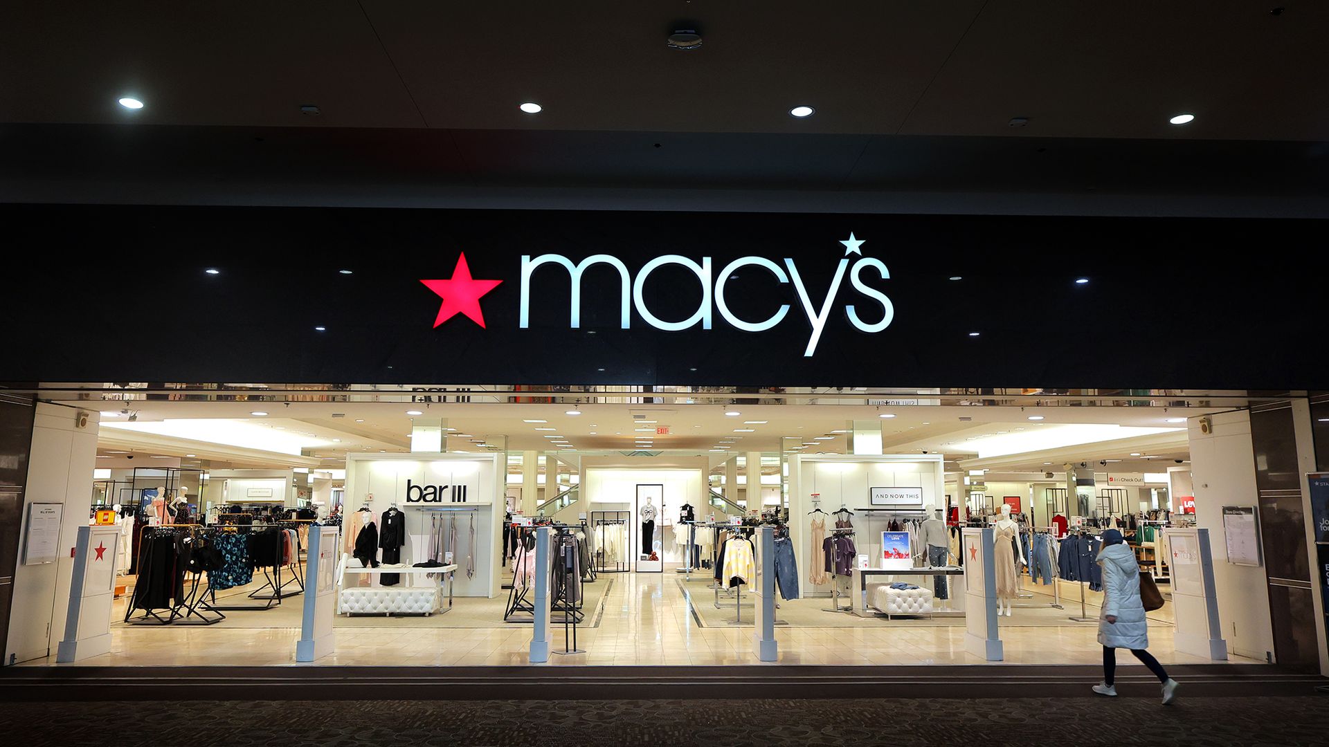 Macy's storefront with a large white macys logo and red star on a black facade; bright interior with clothing racks, mannequins, and a shopper walking by.