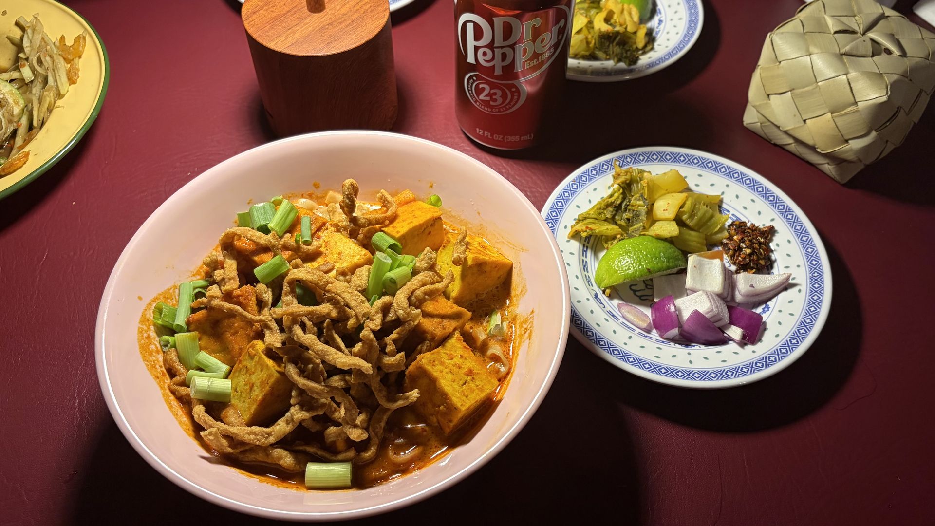 Top-down view of a maroon table: pink bowl of orange curry with tofu and scallions, crispy topping; Dr Pepper can behind; blue-patterned plate with lime and vegetables to the right; woven basket at top right.