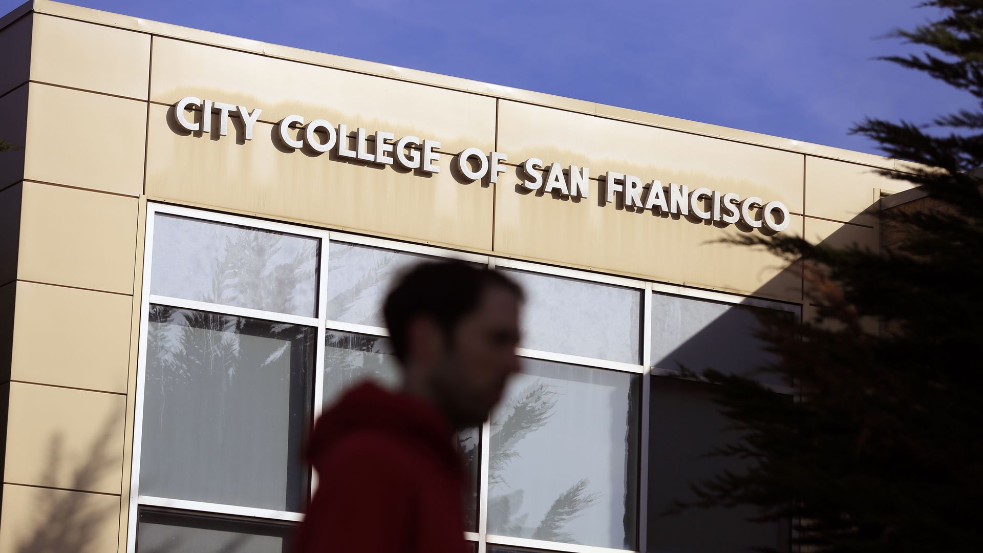 Beige building with large windows and a sign reading City College of San Francisco under a clear blue sky. Silhouette of a person in a red hoodie in front and dark tree branches on the side.