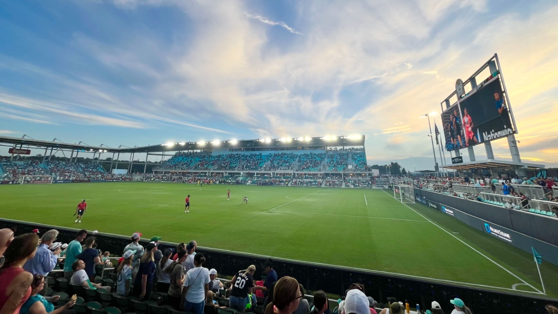 Stadium soccer match at sunset: blue stands filled with fans, bright lights along the upper deck, a large video screen on the right, and players on a green field.