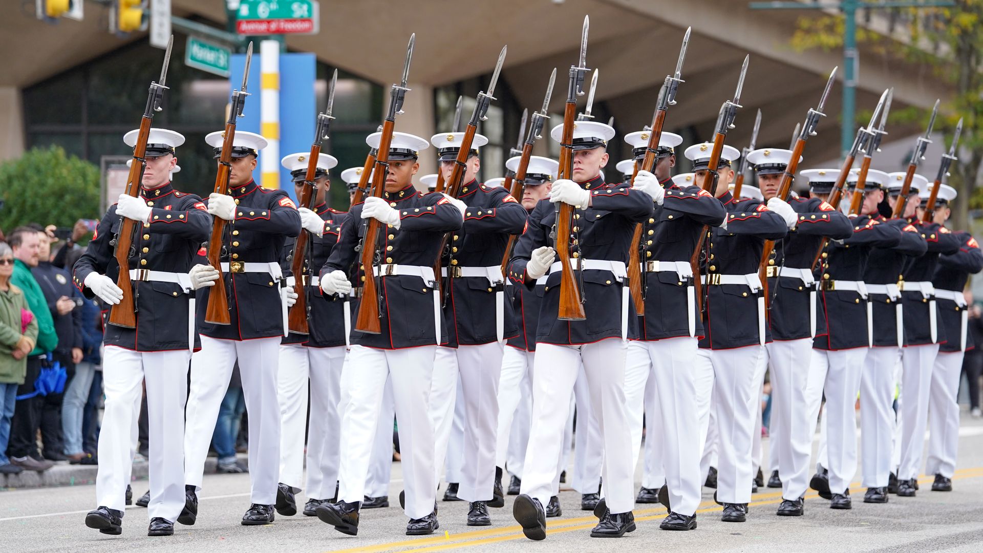 Rows of Marine Corps service members in full uniform marching down a street holding rifles. 