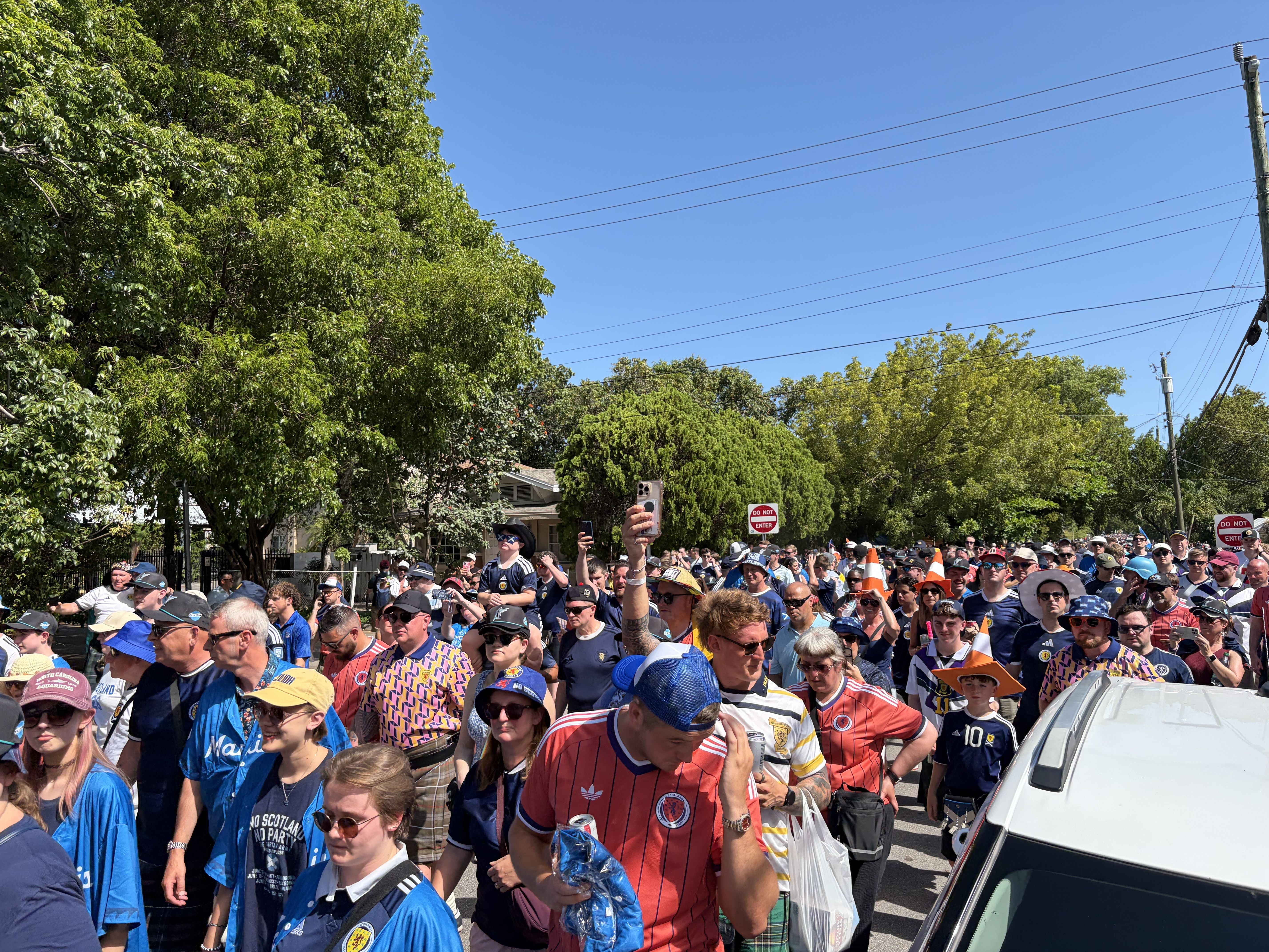 Thousands of fans of Scotland's World Cup team march through Little Haiti on a sunny day, with trees visible in the backround. The fans are in a variety of attire, including jerseys and kilts.