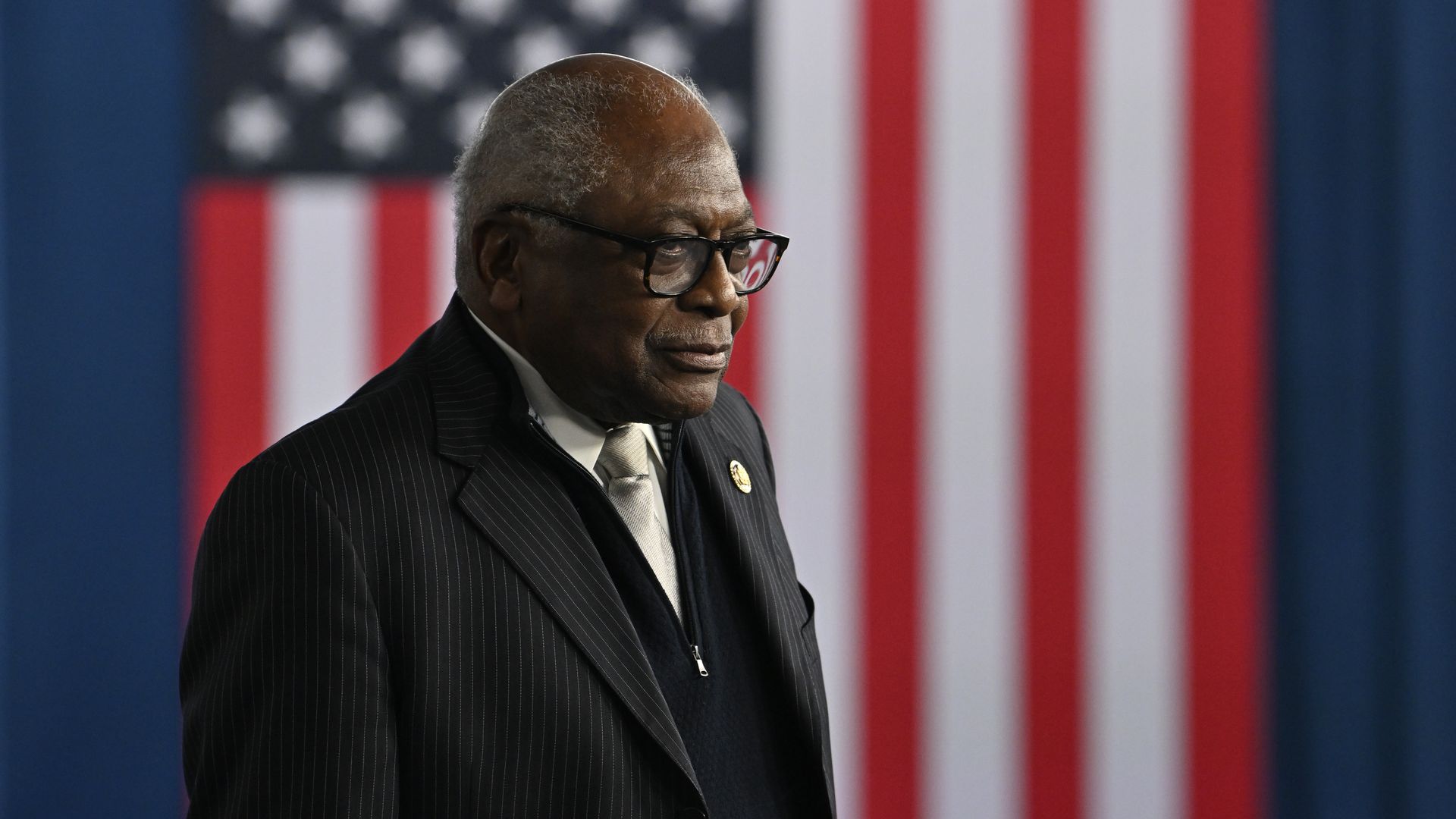 A man in a pinstriped suit and light tie, wearing glasses, stands in front of a large red, white, and blue American flag backdrop.