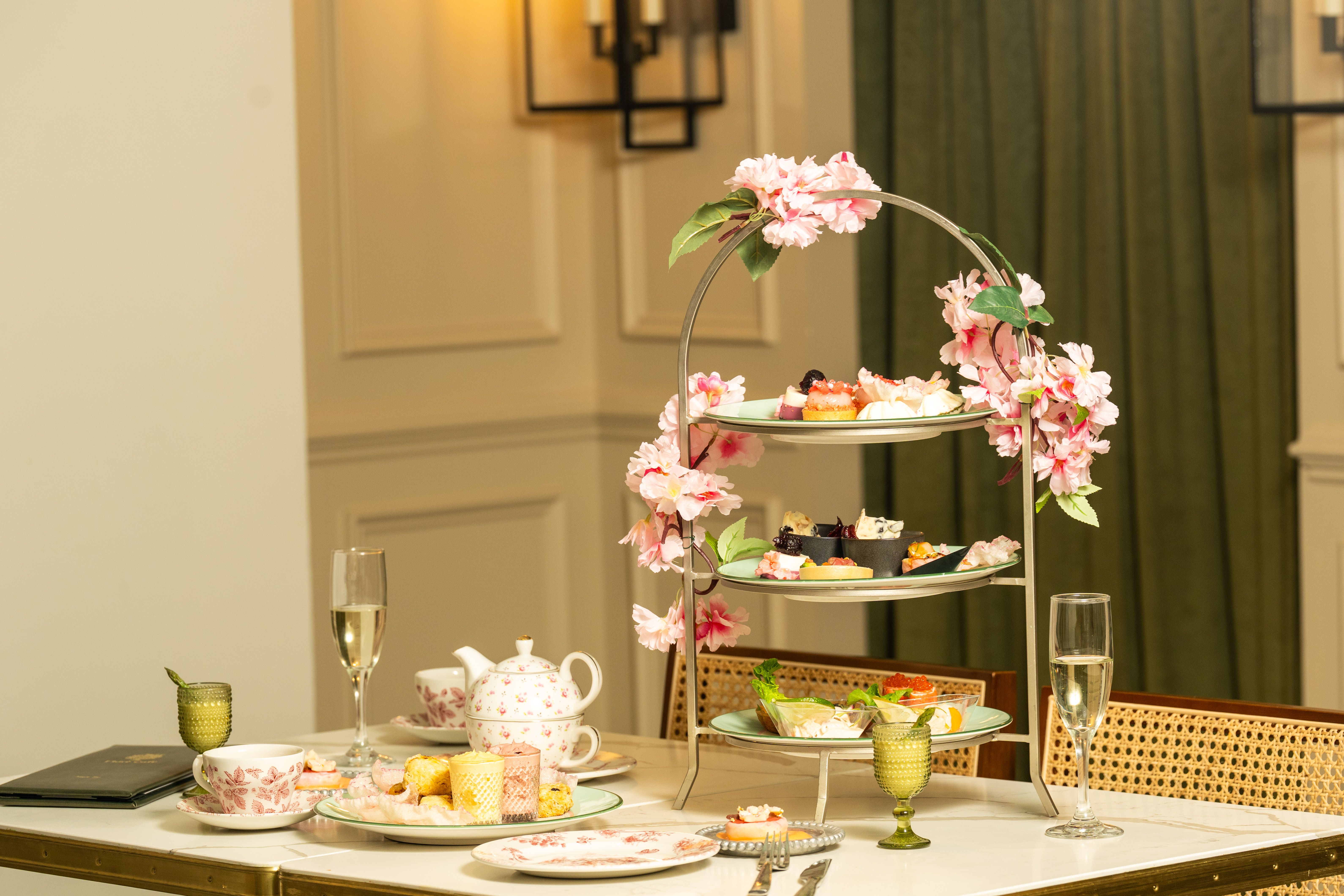 A three-tiered metal stand wrapped with pink flowers holds assorted pastries and savory bites on a marble table; teapot, floral cups, and green goblets add to an elegant tea setup.