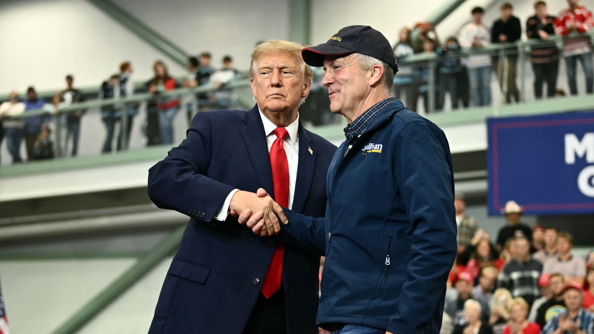 President Trump — wearing a dark blue suit, a red tie, a white collared shirt and an American flag pin — shakes hands with another man wearing jeans and a blue jacket, Sen. Dan Sullivan.
